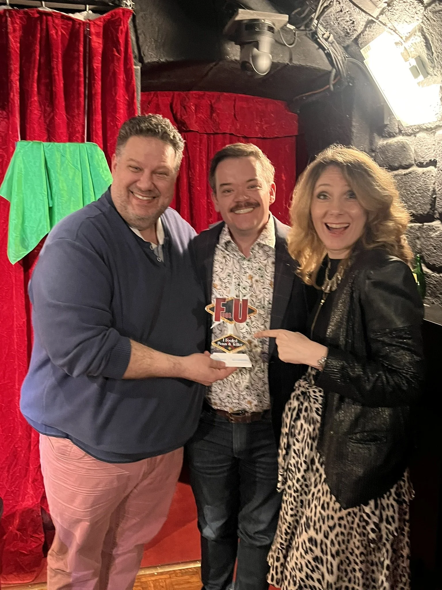 Three people are smiling and posing for a photo at an indoor event with red curtains in the background. The person on the left is a man wearing a dark blue jacket and light-colored pants. The person in the middle is a man with a mustache, wearing a p