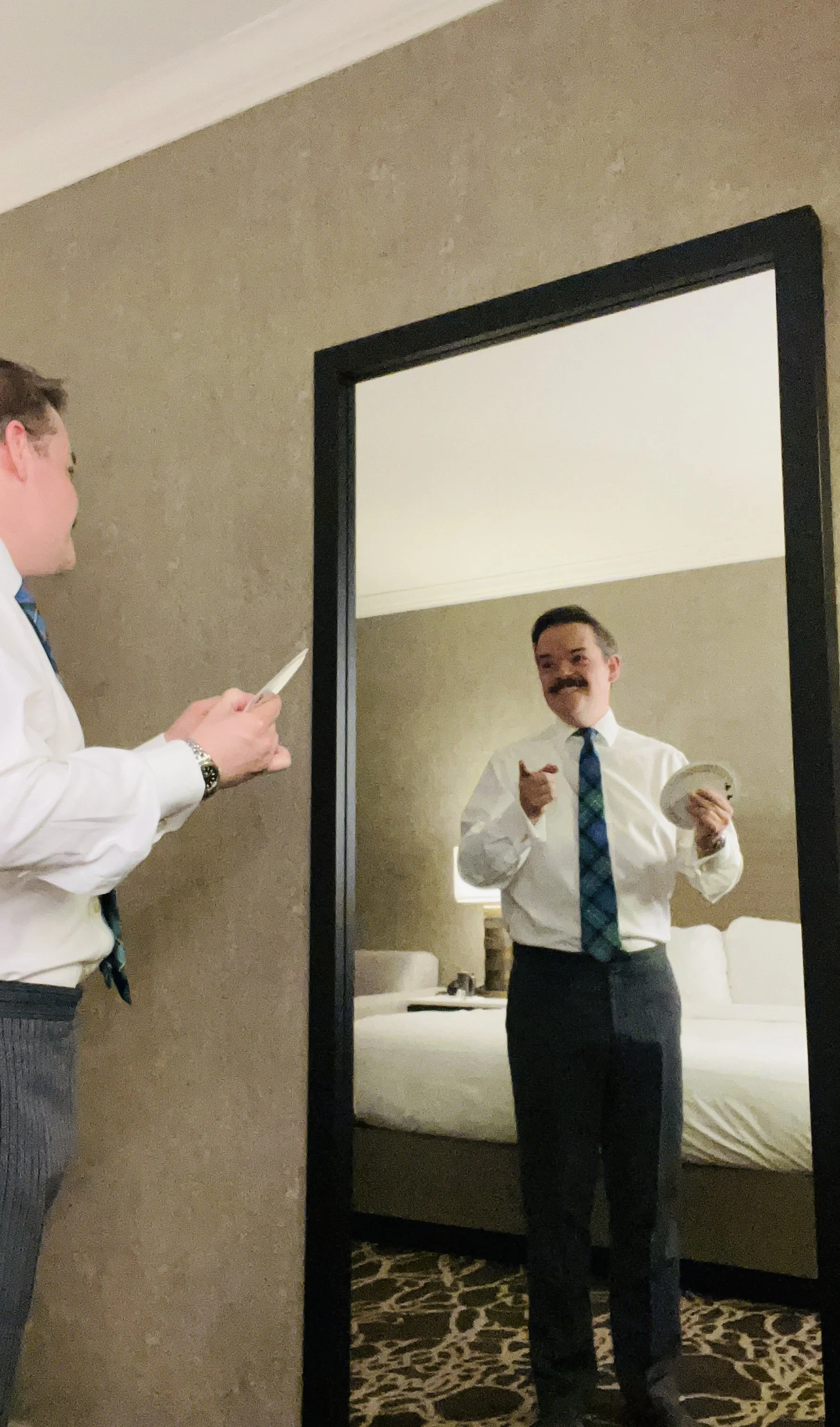 A man with a mustache and dress shirt is looking into a mirror, fanning a deck of cards looking at his reflection. 