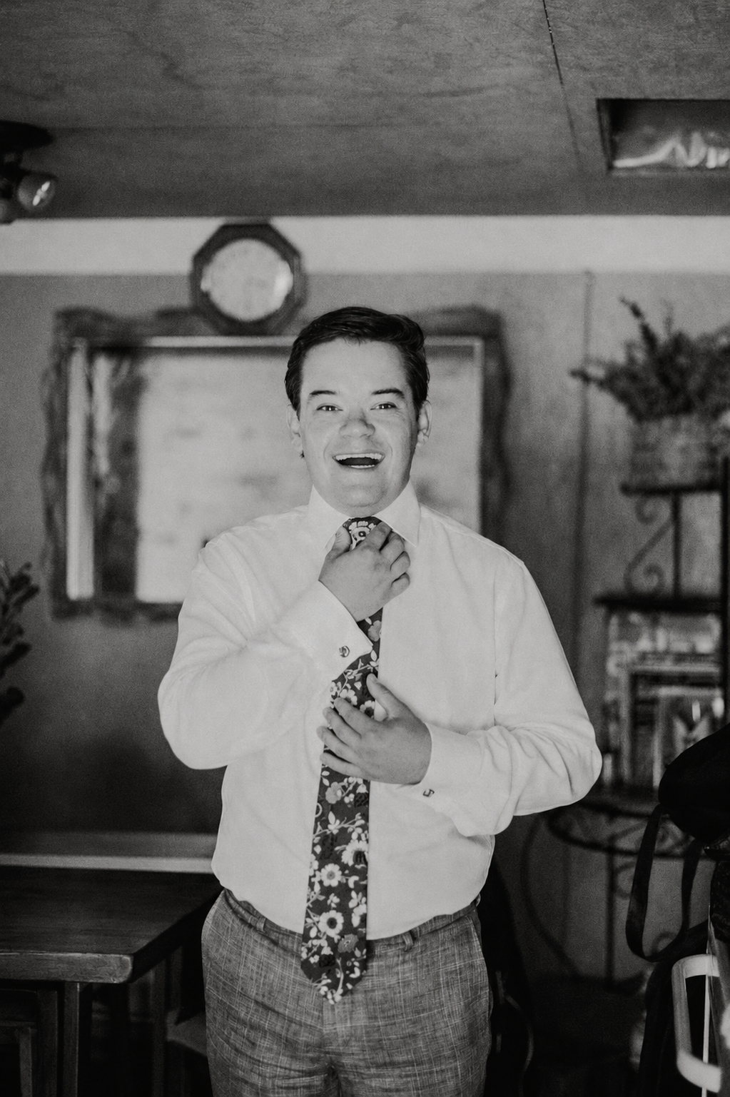 A young man in a white dress shirt and patterned tie, smiling and adjusting his tie, standing in a room with a chalkboard and a clock on the wall.