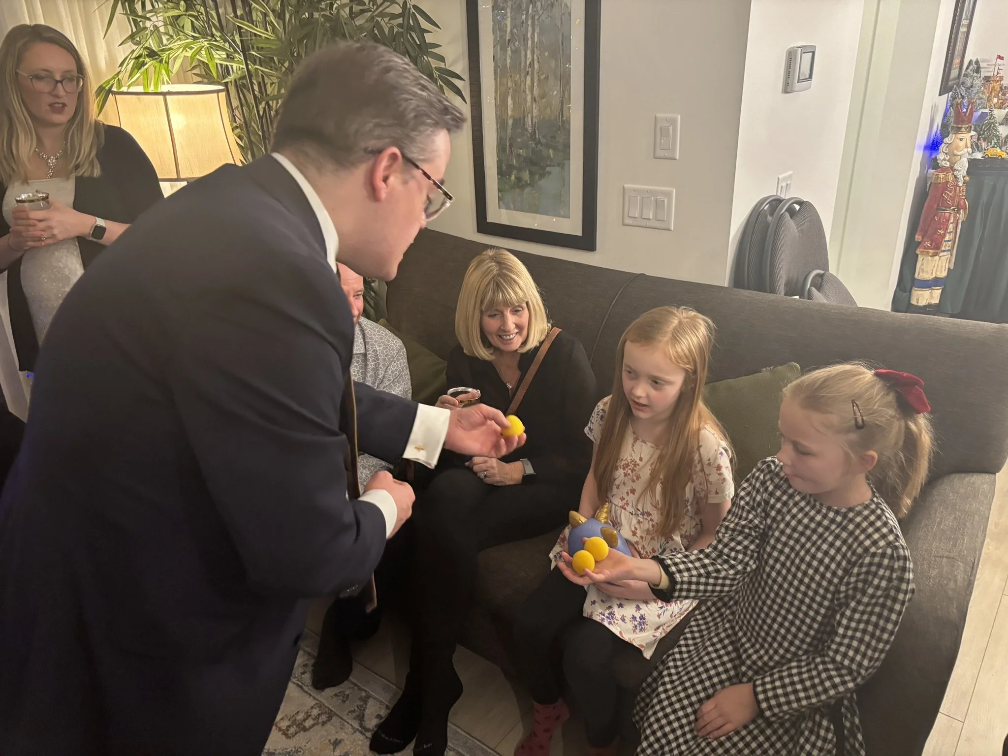 A man in a suit and glasses is handing yellow and purple plastic eggs to young girls at a holiday gathering, with adults and holiday decorations in the background.