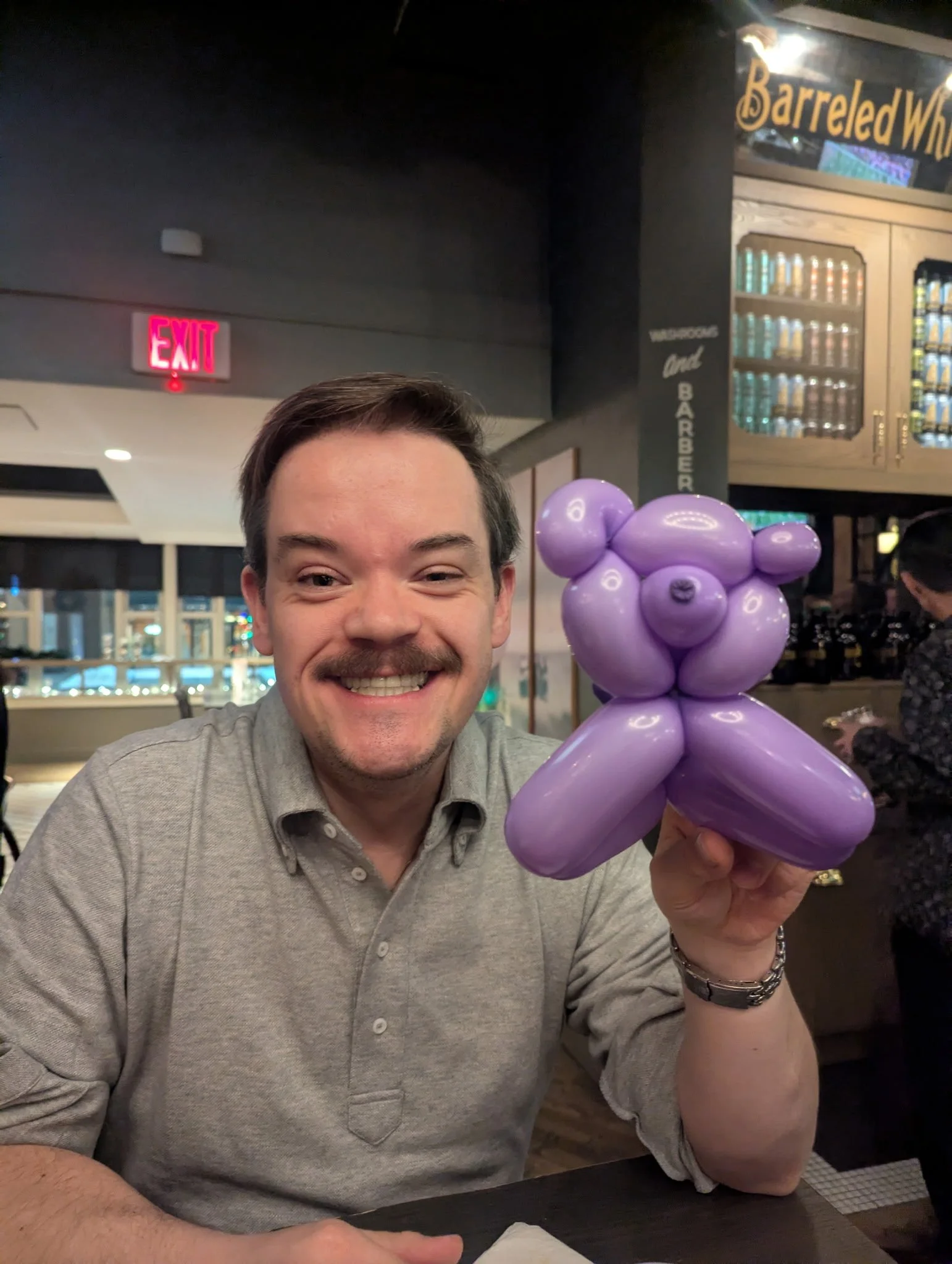 A man in a light gray shirt smiling, holding purple balloon art in the shape of a bear, sitting at a table in a restaurant or bar. There is an exit sign and a bar with drinks in the background.