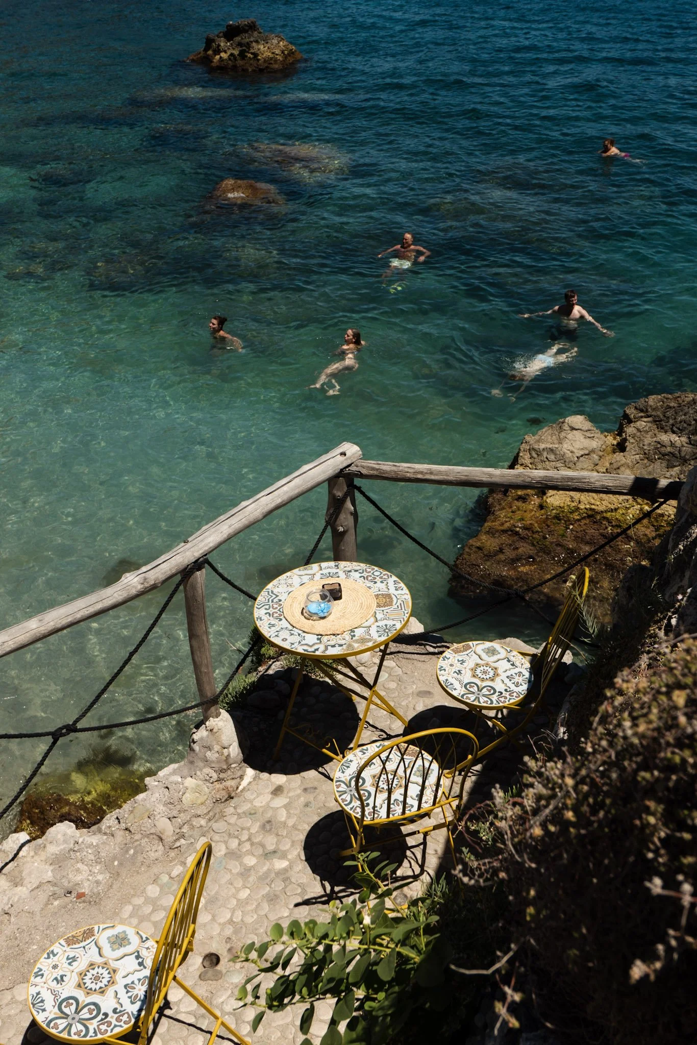 People swimming and relaxing in the clear blue water near rocky shoreline, with a small outdoor seating area with patterned tables and yellow chairs visible in the foreground.