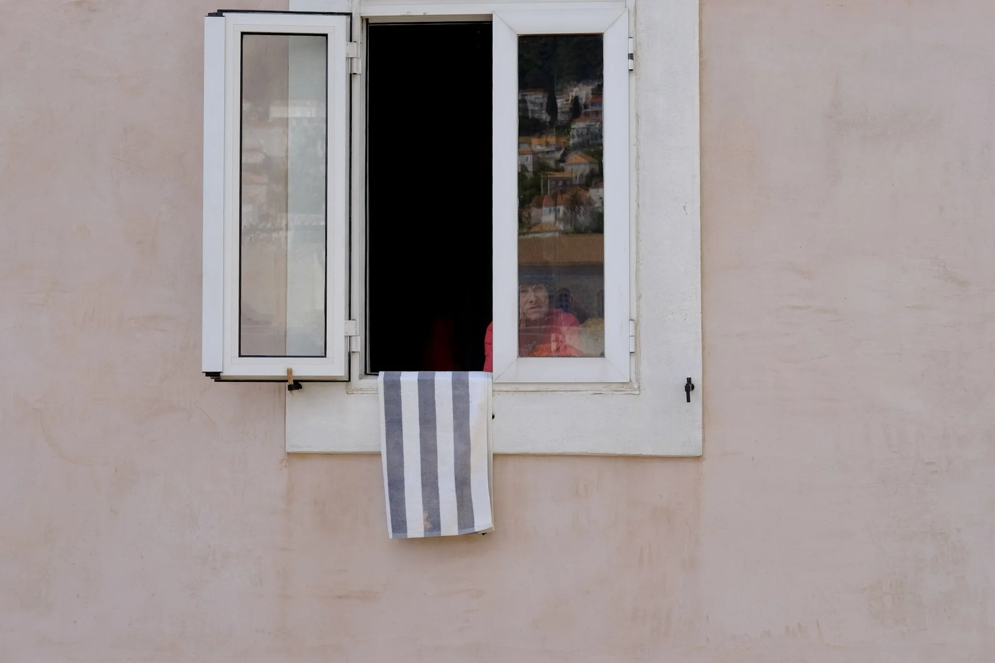 A window with open shutters on a beige wall, with a woman visible inside the room looking outside.