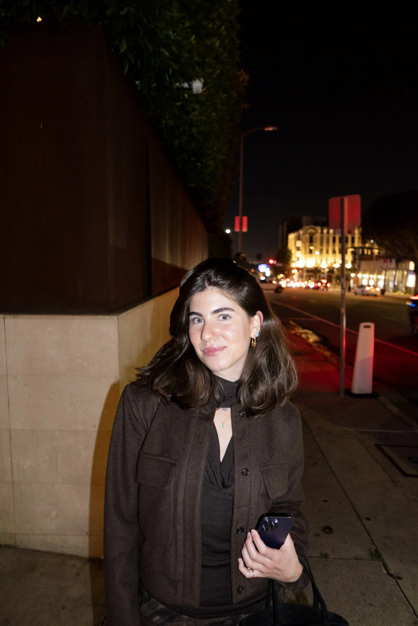 A woman with dark brown hair, blue eyes, and earrings, wearing a dark brown coat and holding a smartphone, standing on a city sidewalk at night with illuminated buildings and streetlights in the background.