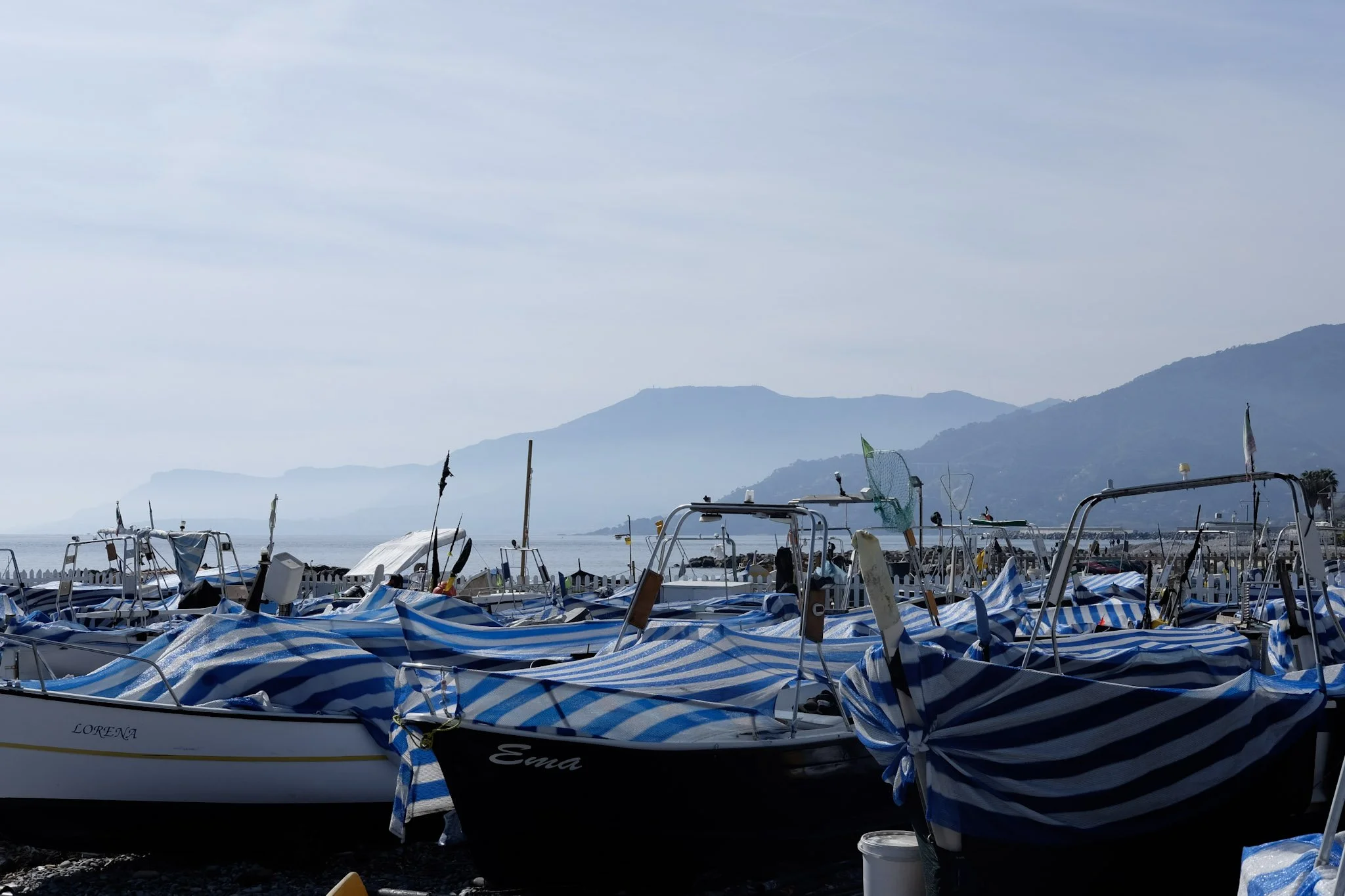 Blick auf zahlreiche Boote am Hafen, einige sind mit blauen und weißen Streifen bedeckt, im Hintergrund Berge und ein bewölkter Himmel.