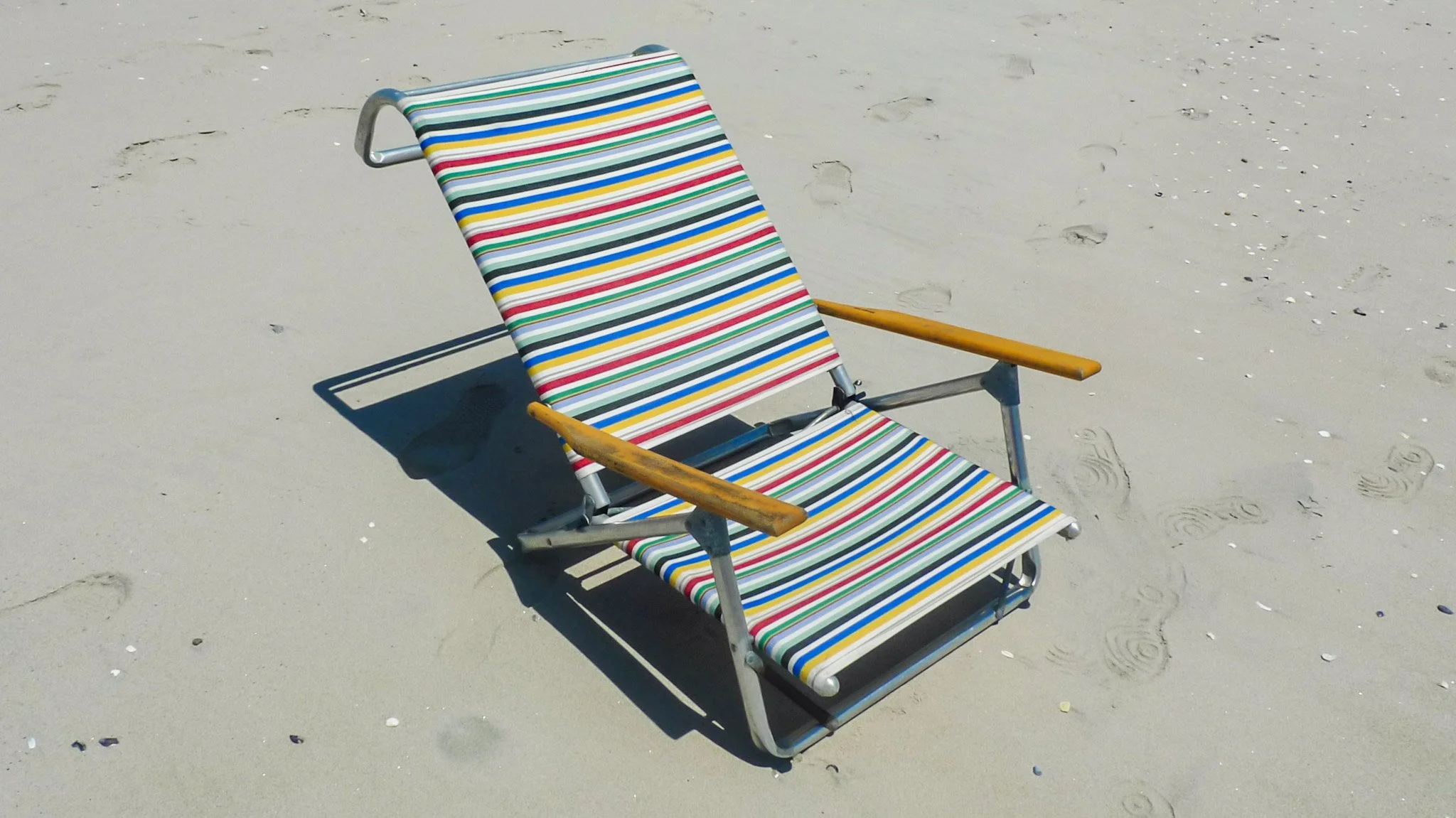 A colorful striped beach chair on sandy beach with footprints and shells.