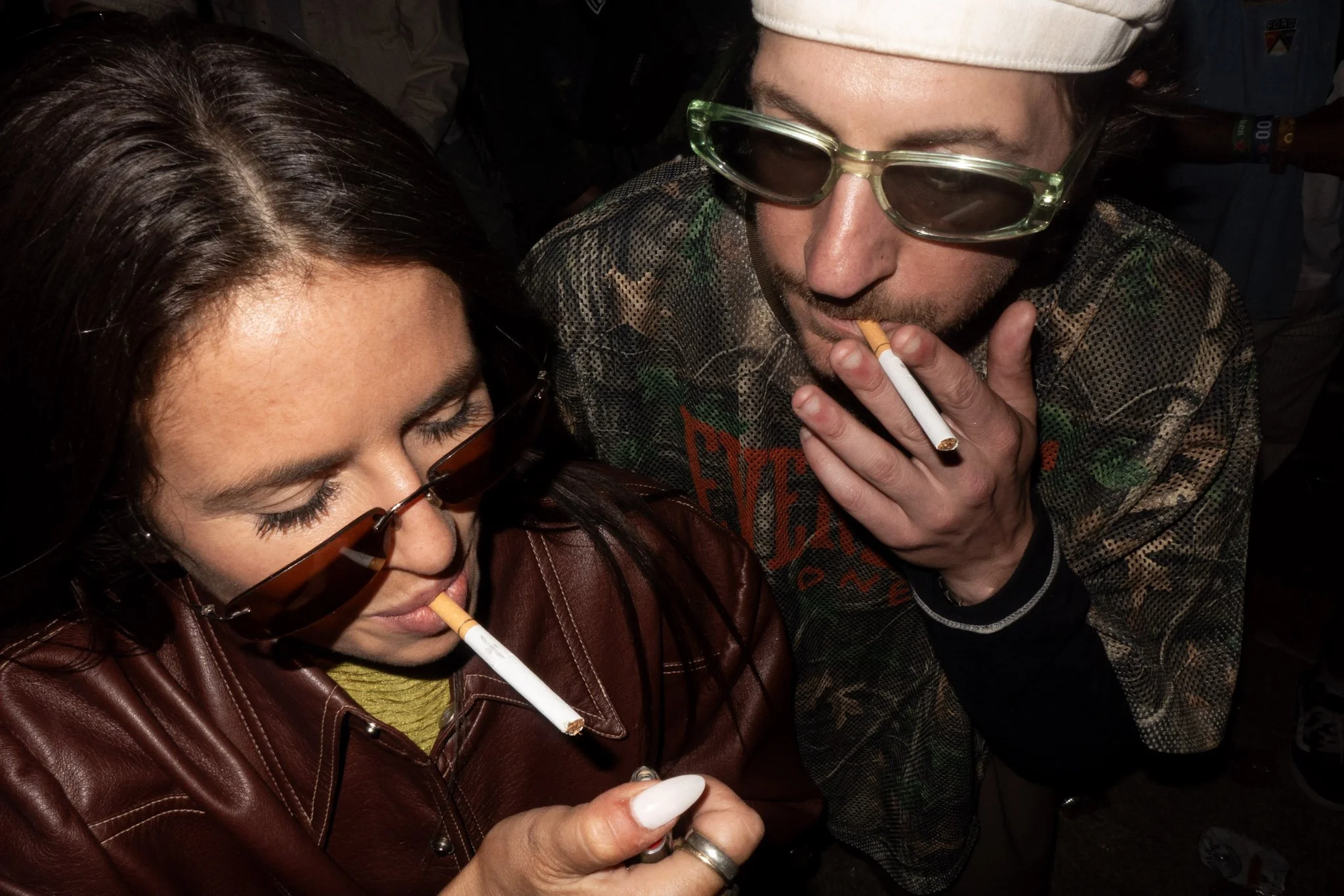 Close-up of a man and a woman smoking cigarettes with cigarettes dipped in white liquid, possibly milk, at a social gathering or party.