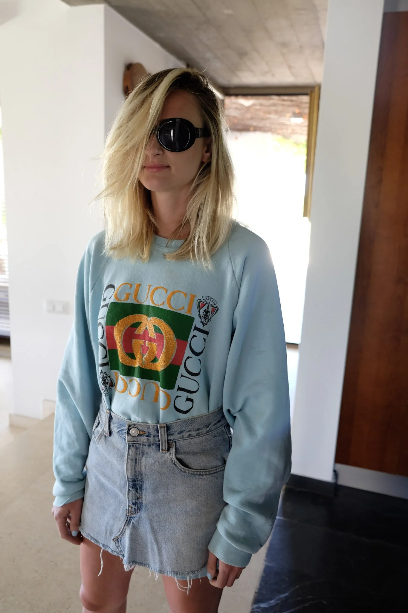A woman with blonde hair wearing sunglasses, a light blue Gucci sweatshirt, and denim shorts standing indoors in front of a mirror.
