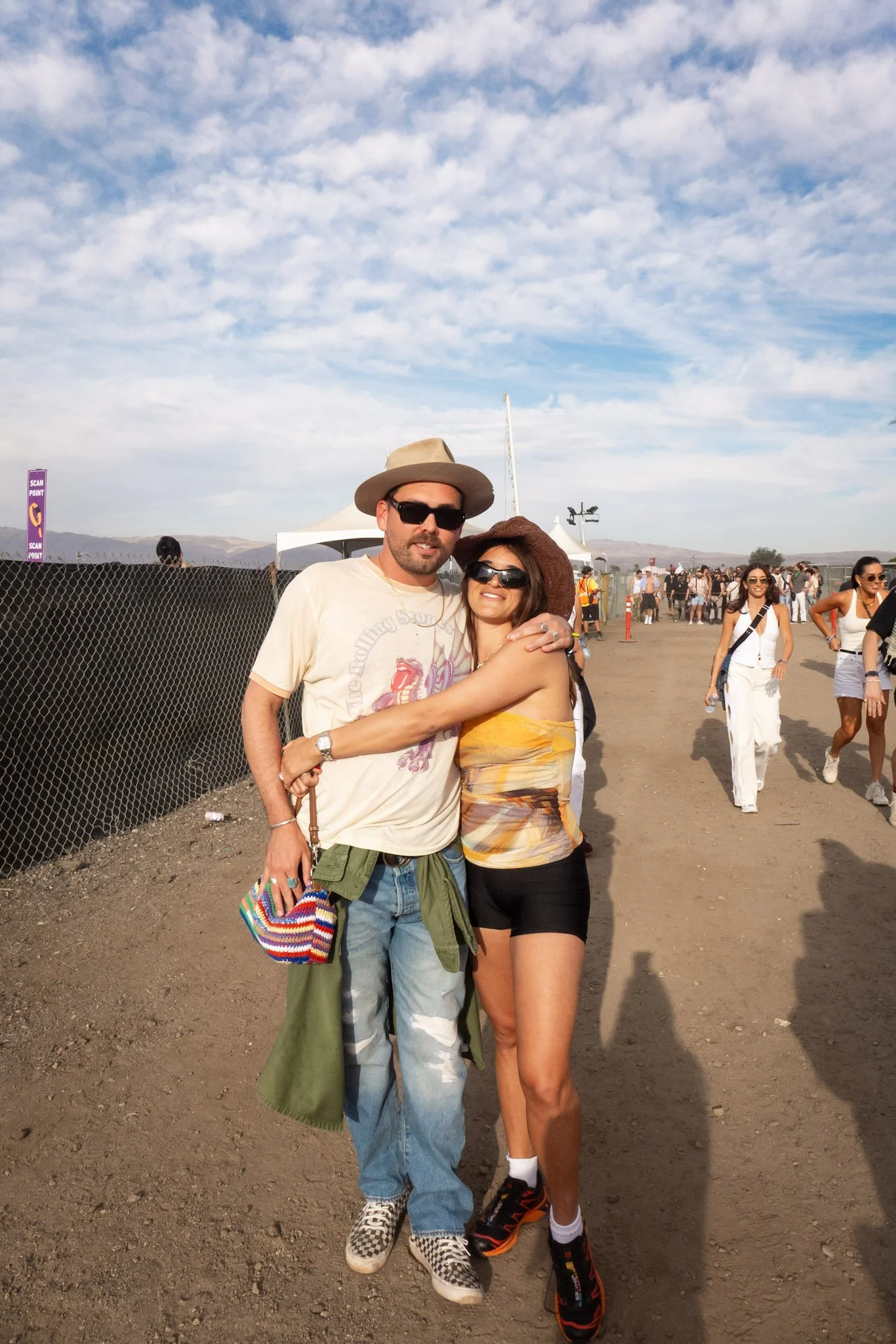 A man and a woman hugging and posing together at a music festival or outdoor event. The man is wearing a wide-brimmed hat, sunglasses, a graphic t-shirt, and jeans, while holding a colorful striped bag. The woman is wearing a brown wide-brimmed hat, 
