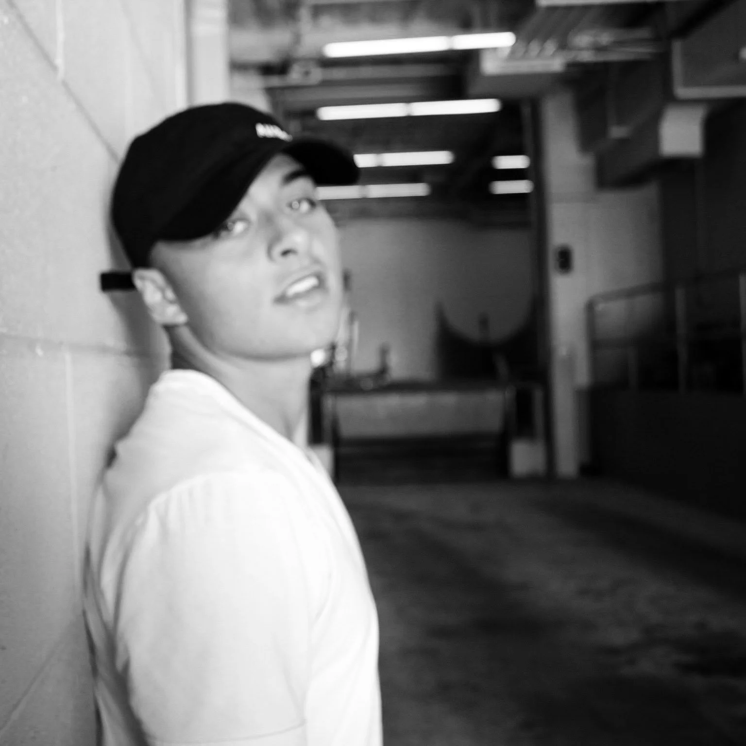 A young person with a cap looks over their shoulder, sitting against a wall in an indoor space with wooden stairs and lighting in the background.