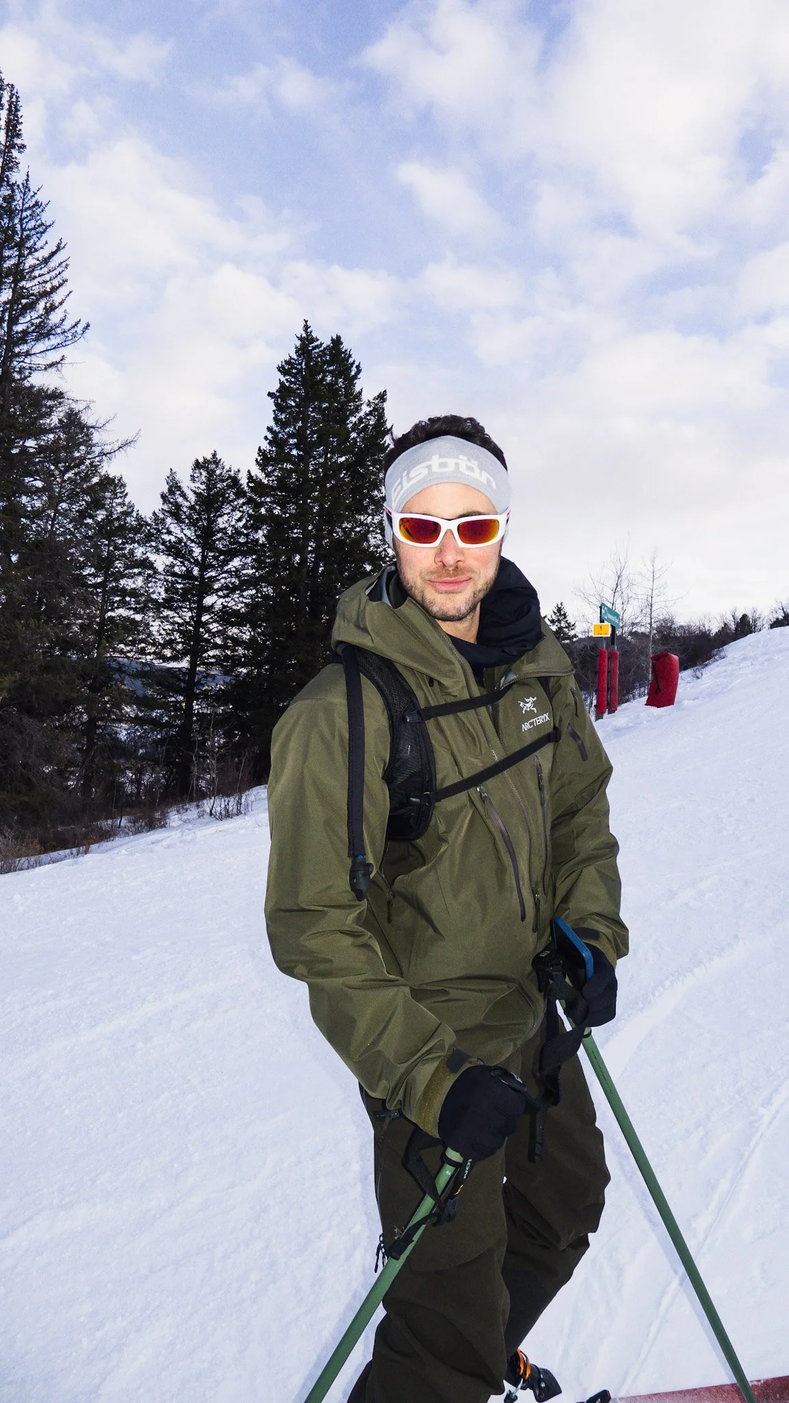 A man dressed in winter gear with sunglasses, standing on snow with trees and a cloudy sky in the background.