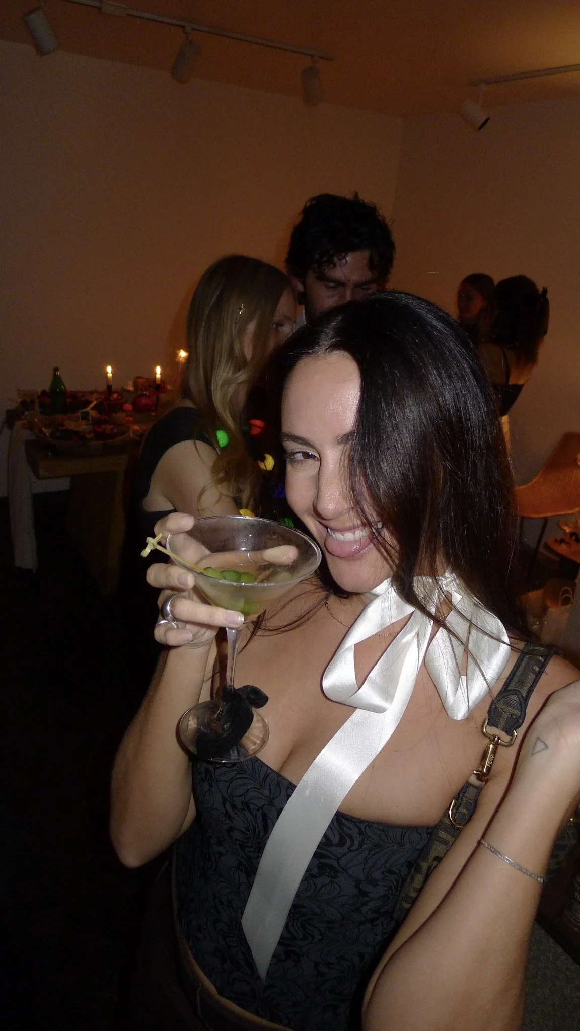 A woman holding a cocktail glass with a lime garnish, smiling, at a party with other people in the background.