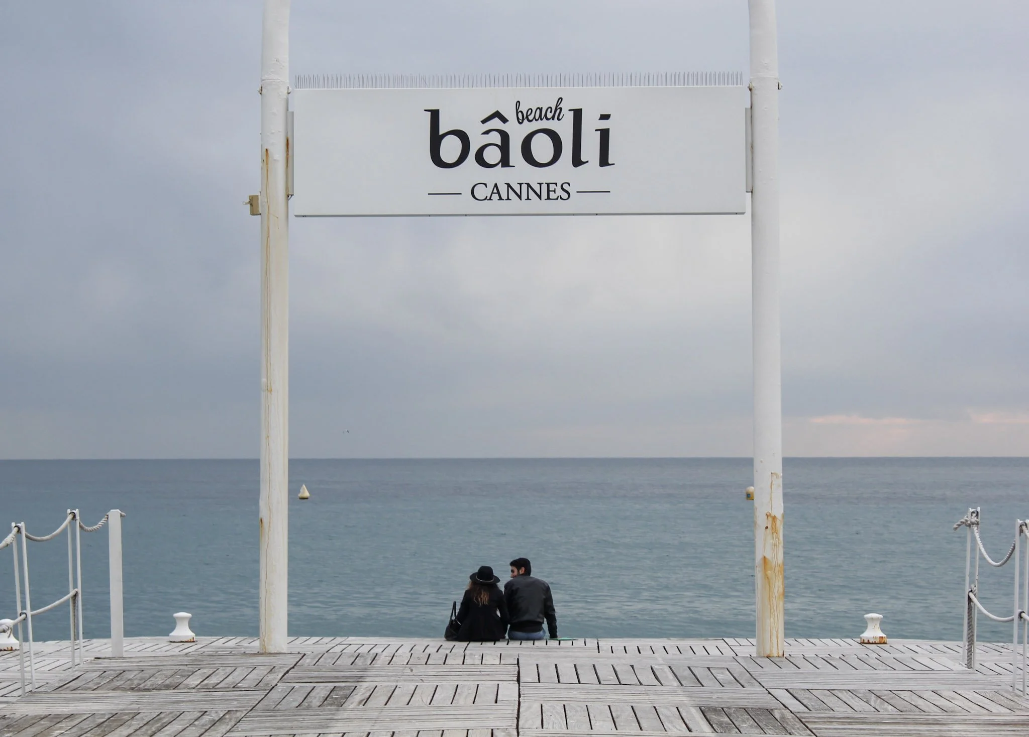 A white signboard on a pier reads 'beach baoli CANNES' with a cloudy sky in the background. Two people sit on the pier facing the sea, with ropes along the sides of the pier.