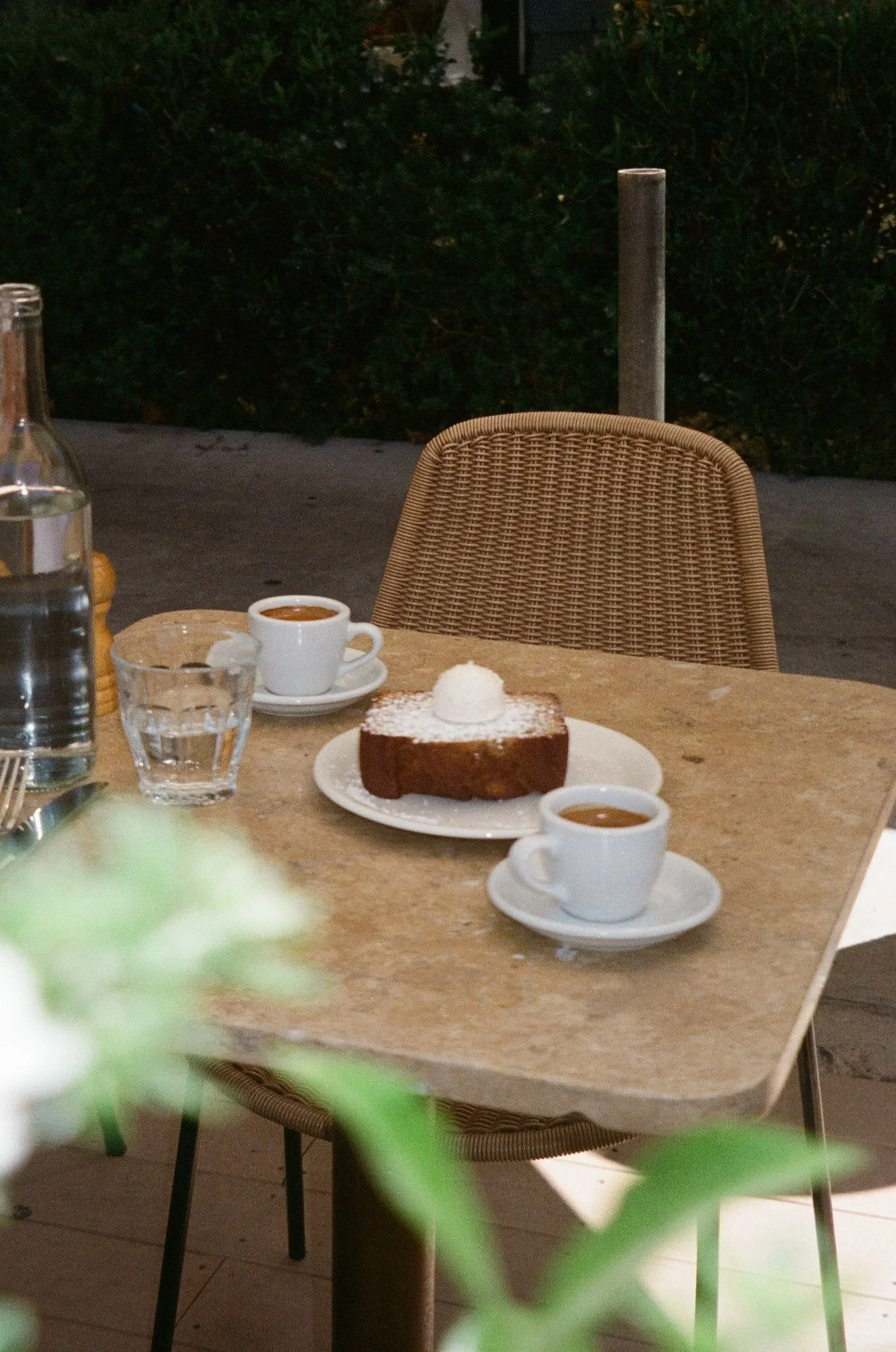 A wooden table with a slice of chocolate cake topped with whipped cream, two cups of coffee, a glass of water, and a bottle of water in the background. There are woven chairs and greenery outside.
