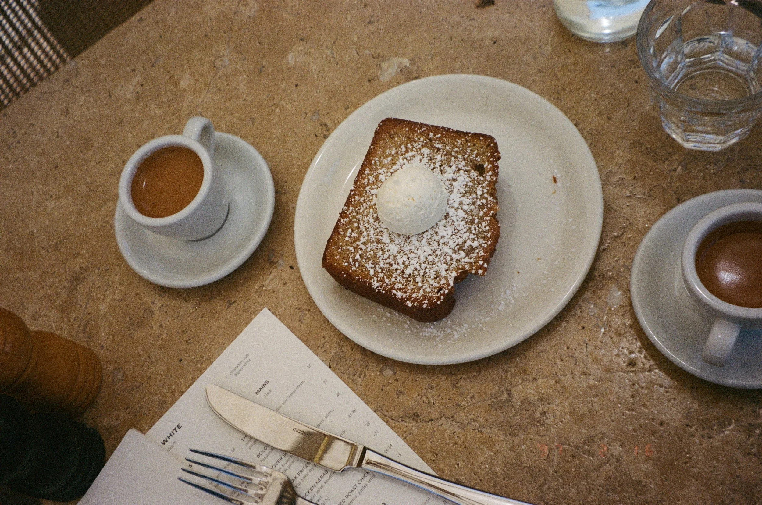 A dessert plate featuring a large piece of toasted bread or pastry, dusted with powdered sugar and topped with a scoop of vanilla ice cream, accompanied by cups of coffee or espresso, on a table with a glass of water, a menu, and utensils.