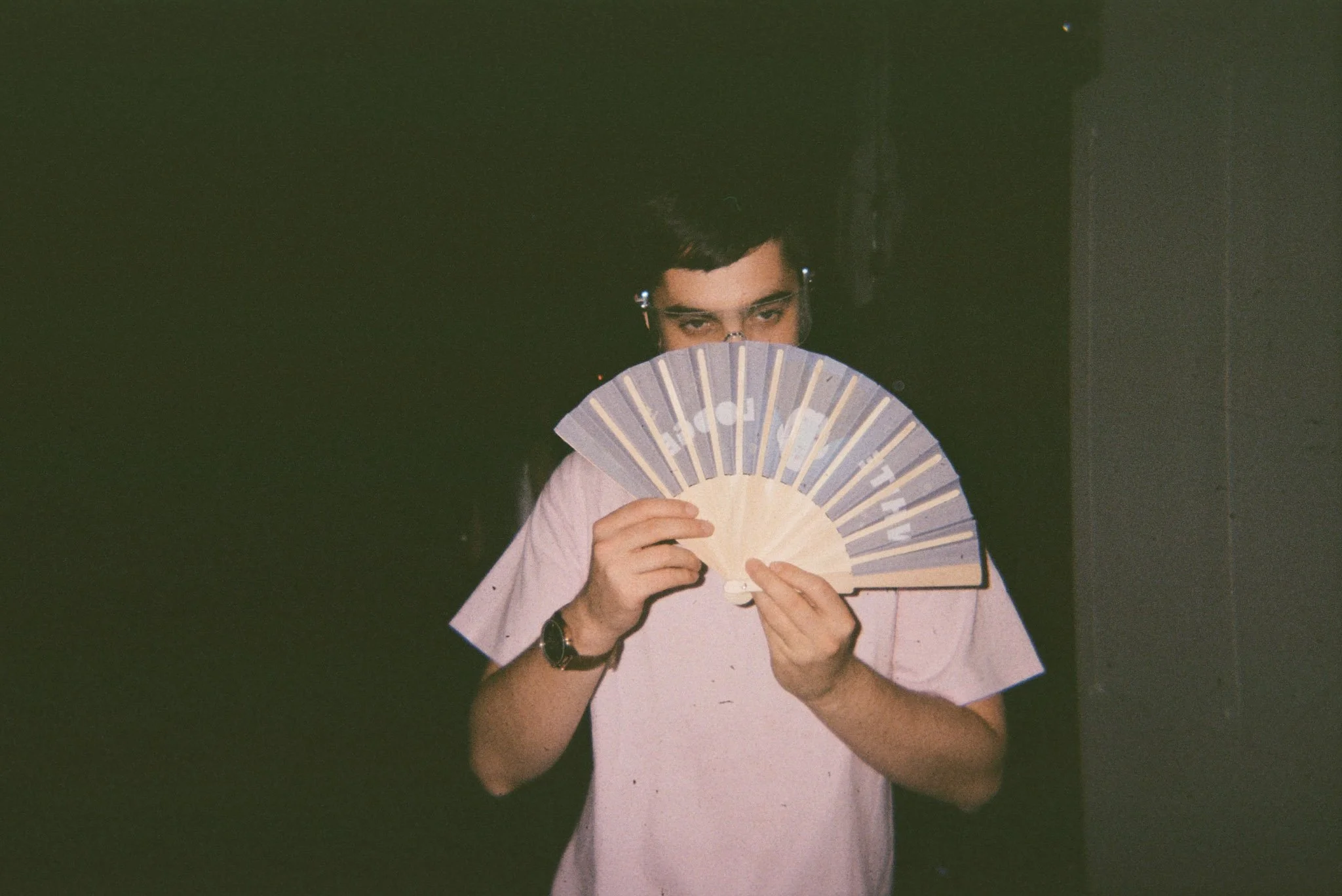 A person with dark hair and glasses holding a closed decorative fan, partially covering their face, with a dark background.