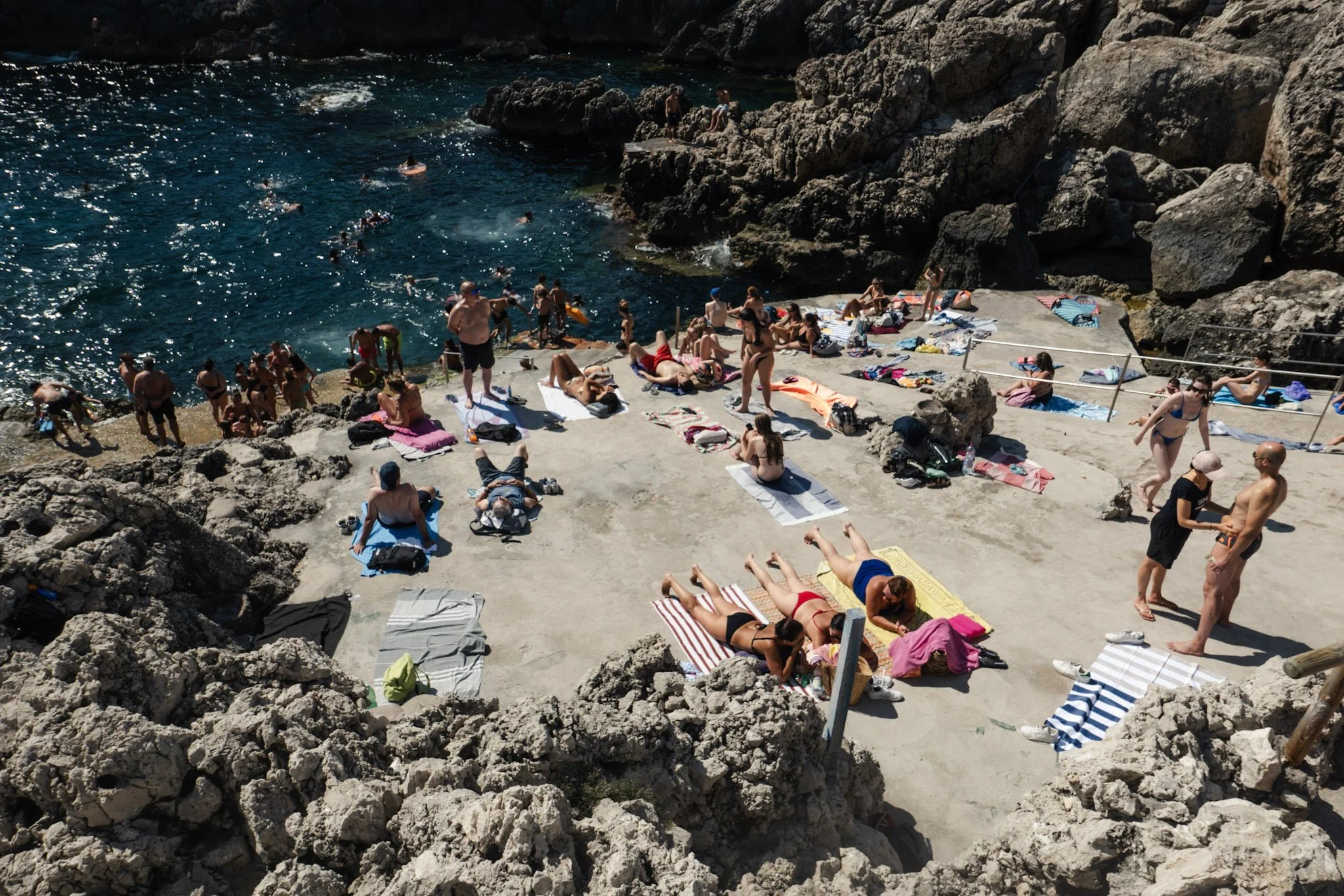 People relaxing on a rocky beach by the ocean, some lying on towels, others sitting or standing, and swimmers in the water, surrounded by large rocks and cliffs under bright sunlight.