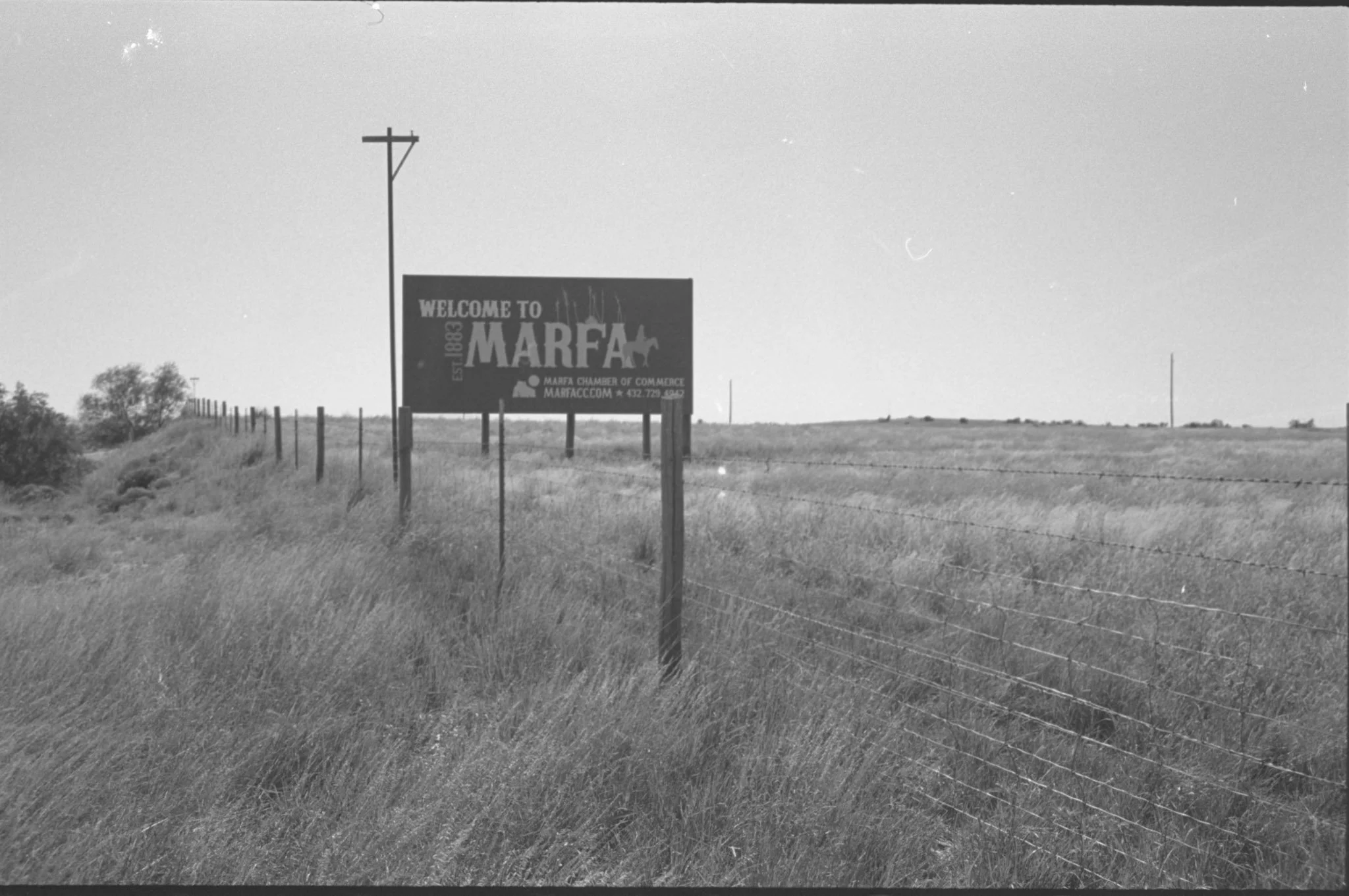 A roadside sign welcoming visitors to Marfa, Texas, with a logo of a horse and rider, and contact information for the Marfa Chamber of Commerce, set against a flat open landscape with a barbed wire fence and utility poles.