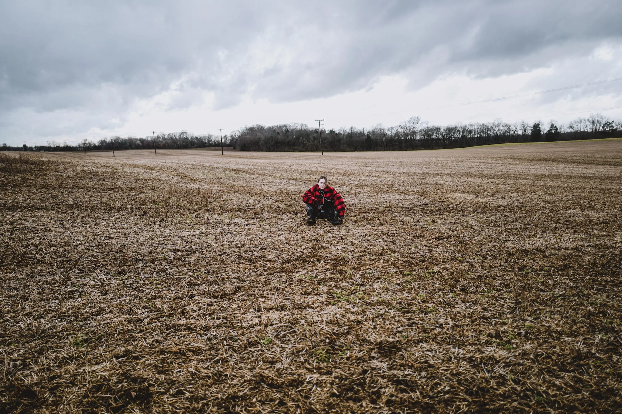 A person wearing a red and black checkered jacket crouching in a wide, open, plowed field under a cloudy sky with trees and power lines in the background.