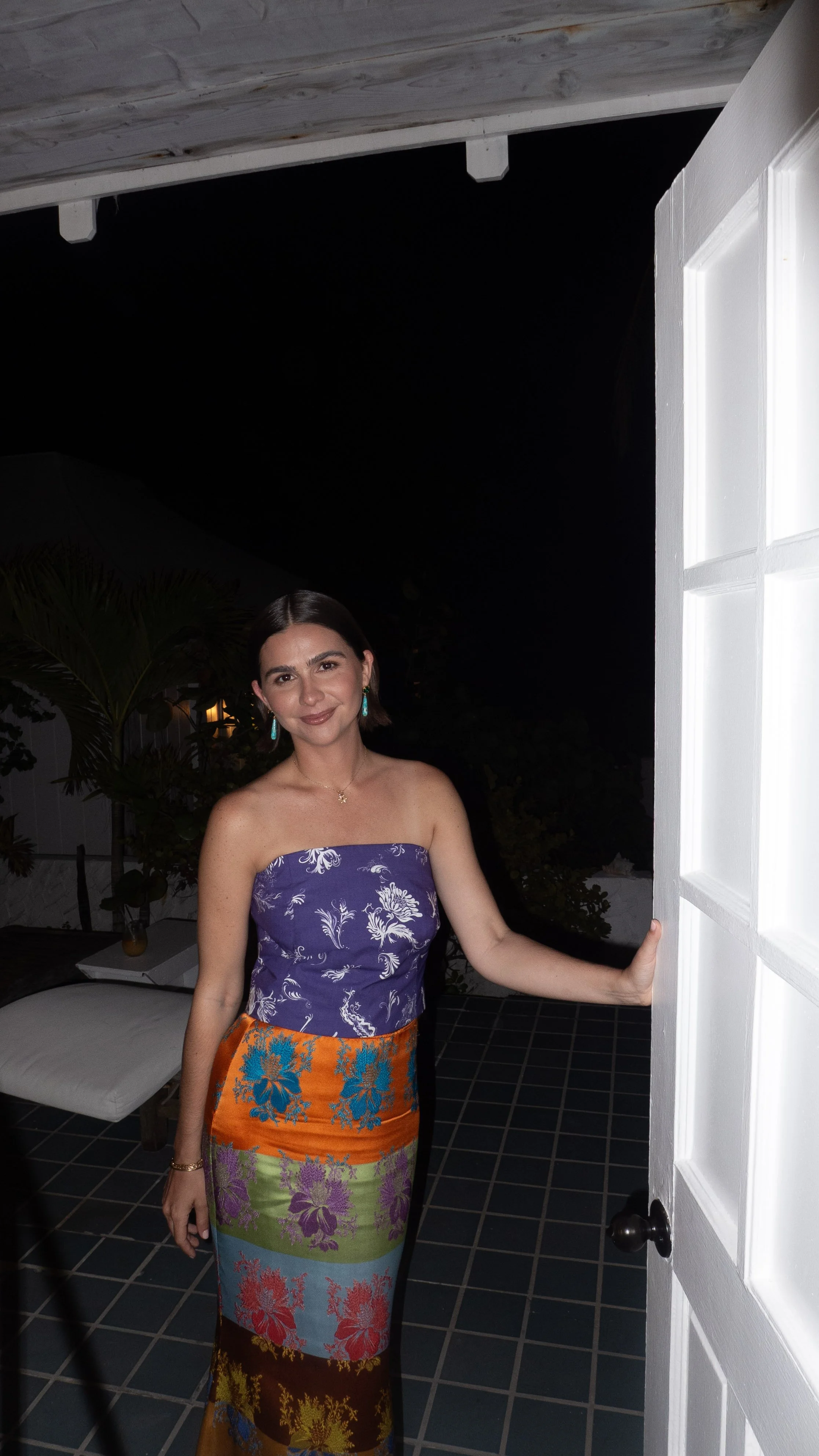 A woman standing at a door opening, wearing a colorful strapless dress with floral patterns and turquoise earrings, at night with dark background and some plants visible behind her.