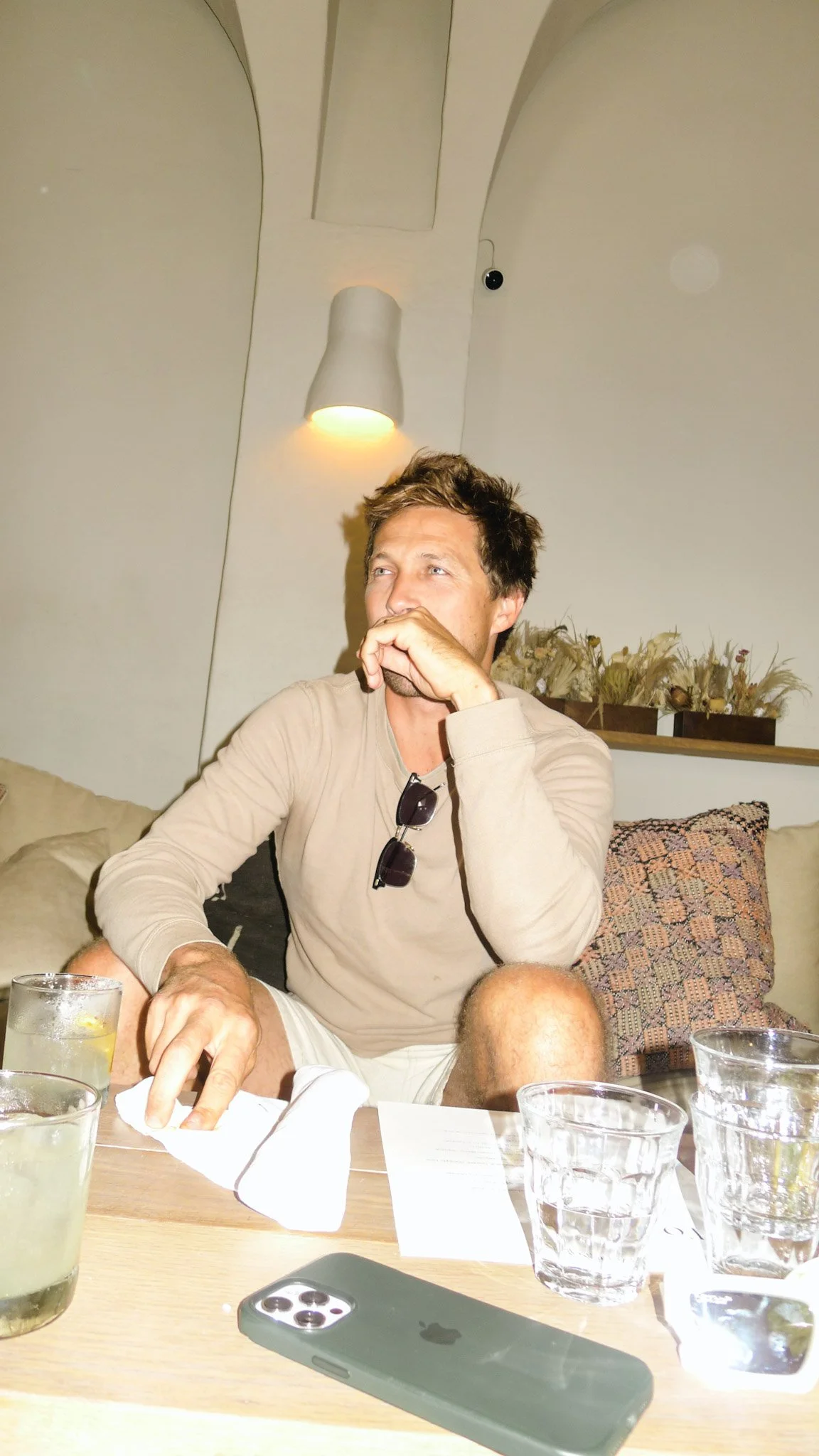 A man sitting at a table with multiple glasses of water, some with lemon slices, in a room with beige walls and a decorative pillow on a couch behind him. He is wearing a beige long-sleeve shirt and white shorts, with sunglasses hanging from his shir