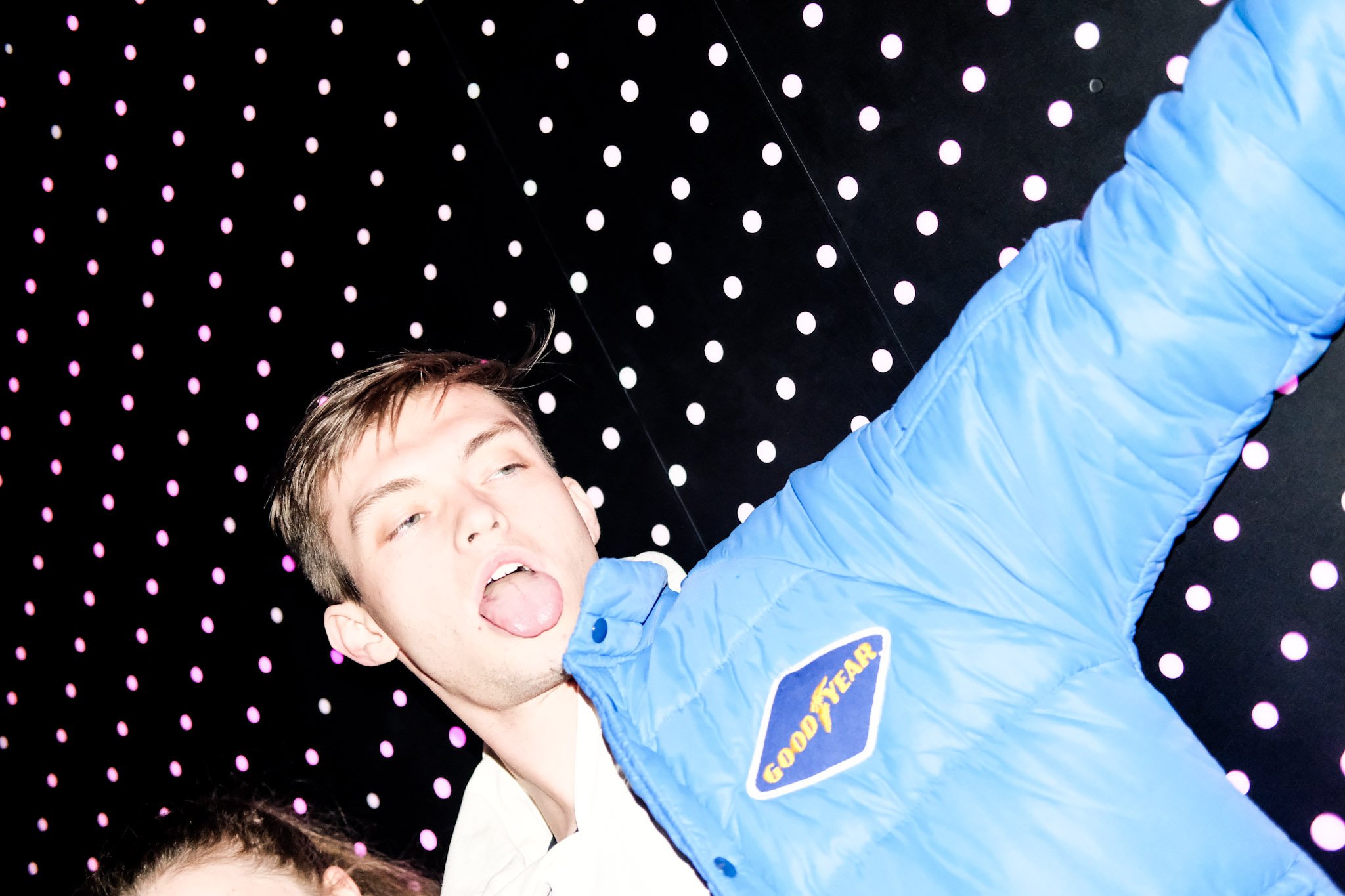 A young person with light skin and brown hair sticking their tongue out, wearing a blue jacket with a logo, in front of a backdrop with pink and white dots.