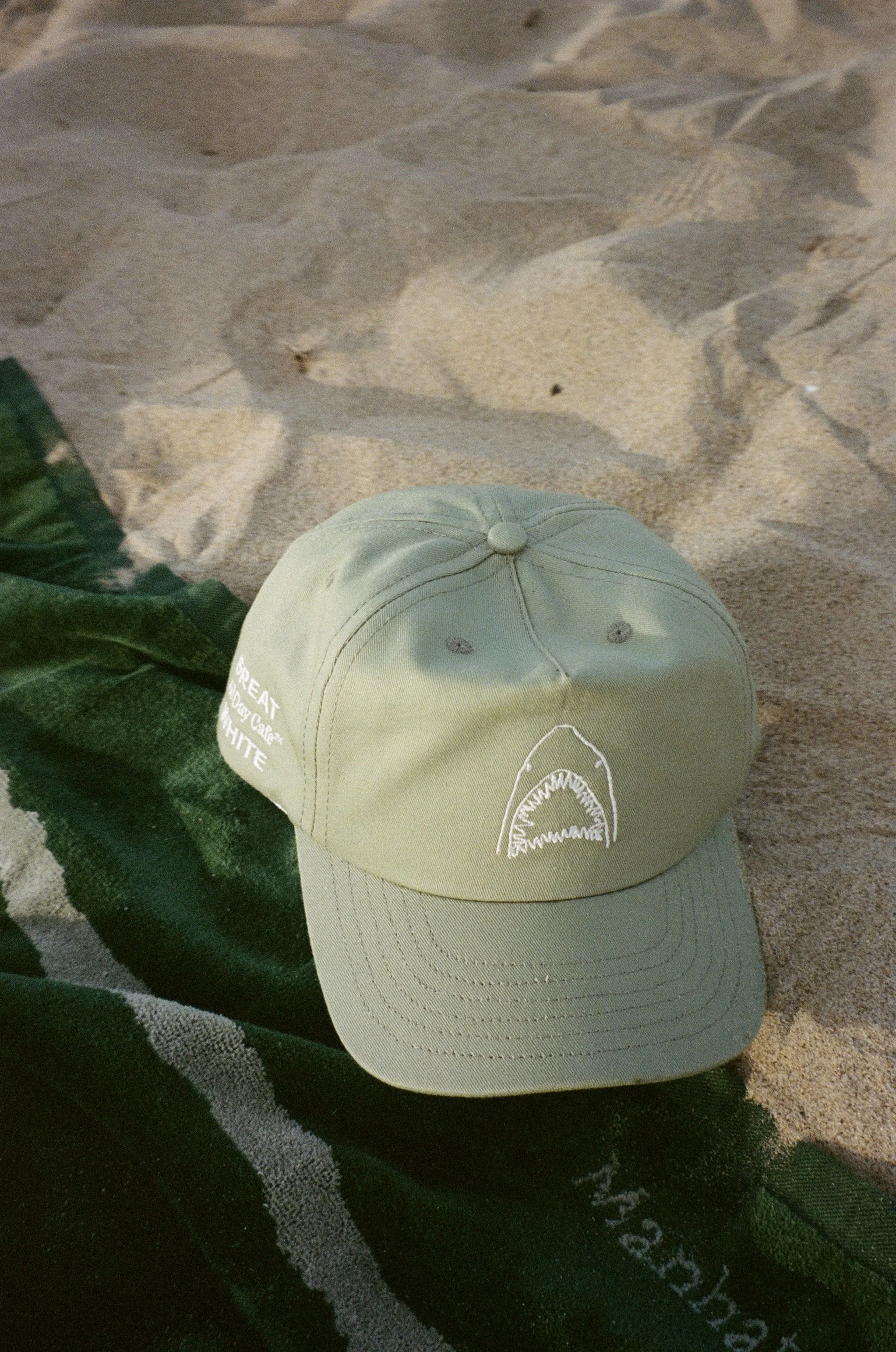 A beige baseball cap with a small logo on the front, placed on a black and gray patterned fabric, with a green object partially visible on the side, on a sandy surface.