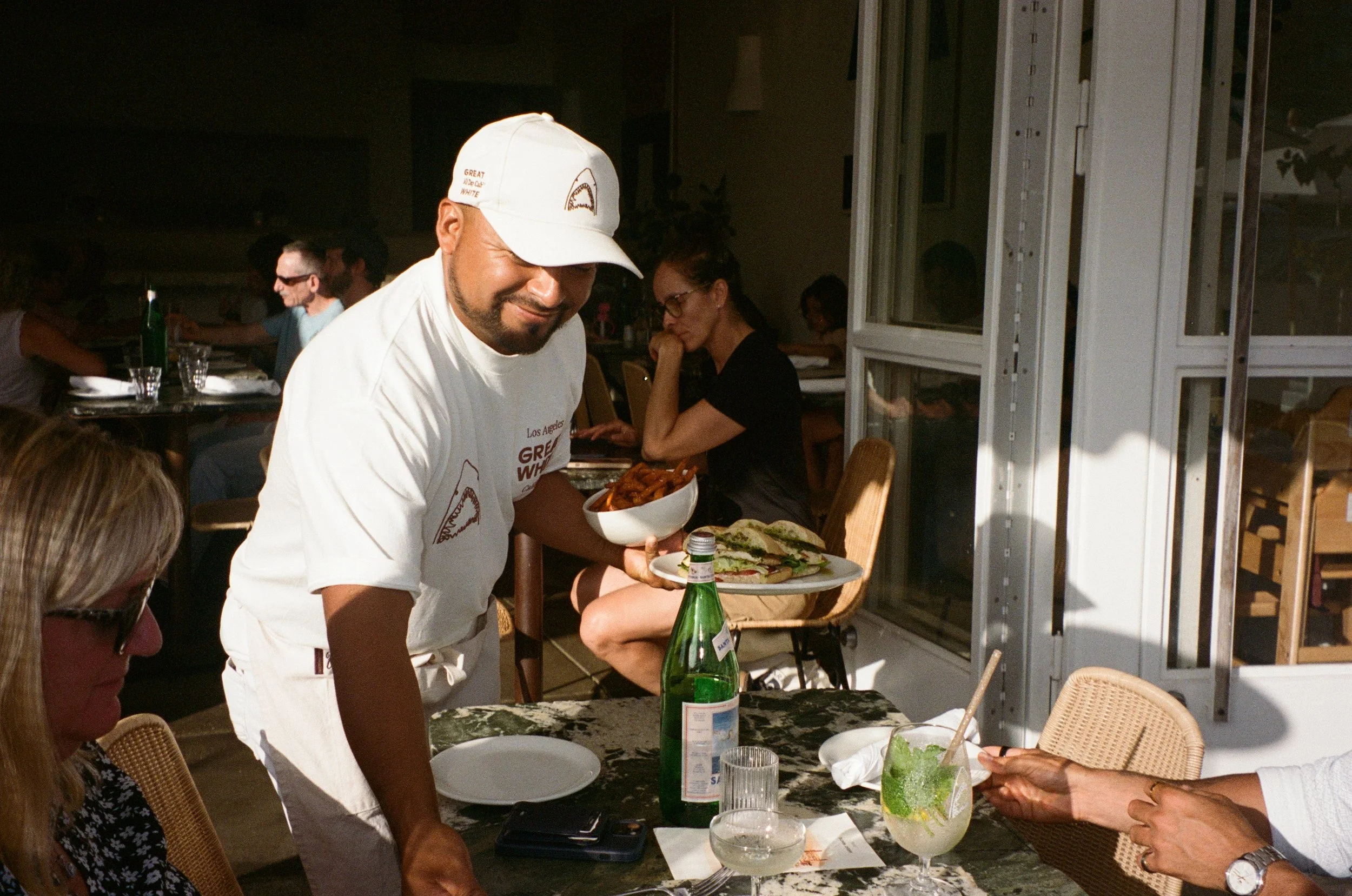 A man wearing a white baseball cap and white T-shirt serving a bowl of food at a dinner party, with guests seated around a table with drinks and food, in a well-lit space with large glass windows.