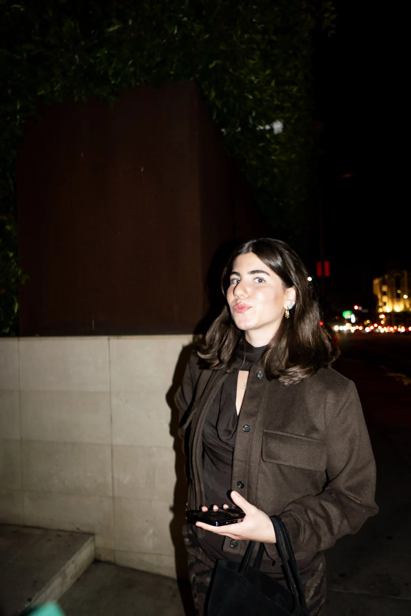 A young woman with dark hair and light skin standing outdoors at night, holding a phone and a black handbag, wearing a brown jacket and earrings, with city lights in the background.