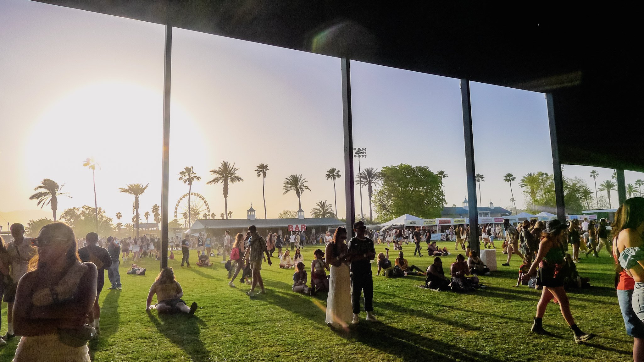 Crowd of people at an outdoor event with palm trees, a Ferris wheel, and food stands, viewed through large window panels at sunset.