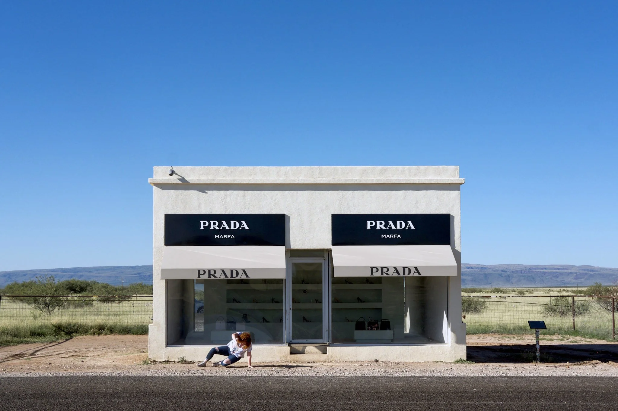 A small Prada storefront with signage and awnings, situated in a rural area under a clear blue sky. A person with curly hair, wearing a white shirt and distressed jeans, is sitting on the ground outside the store.