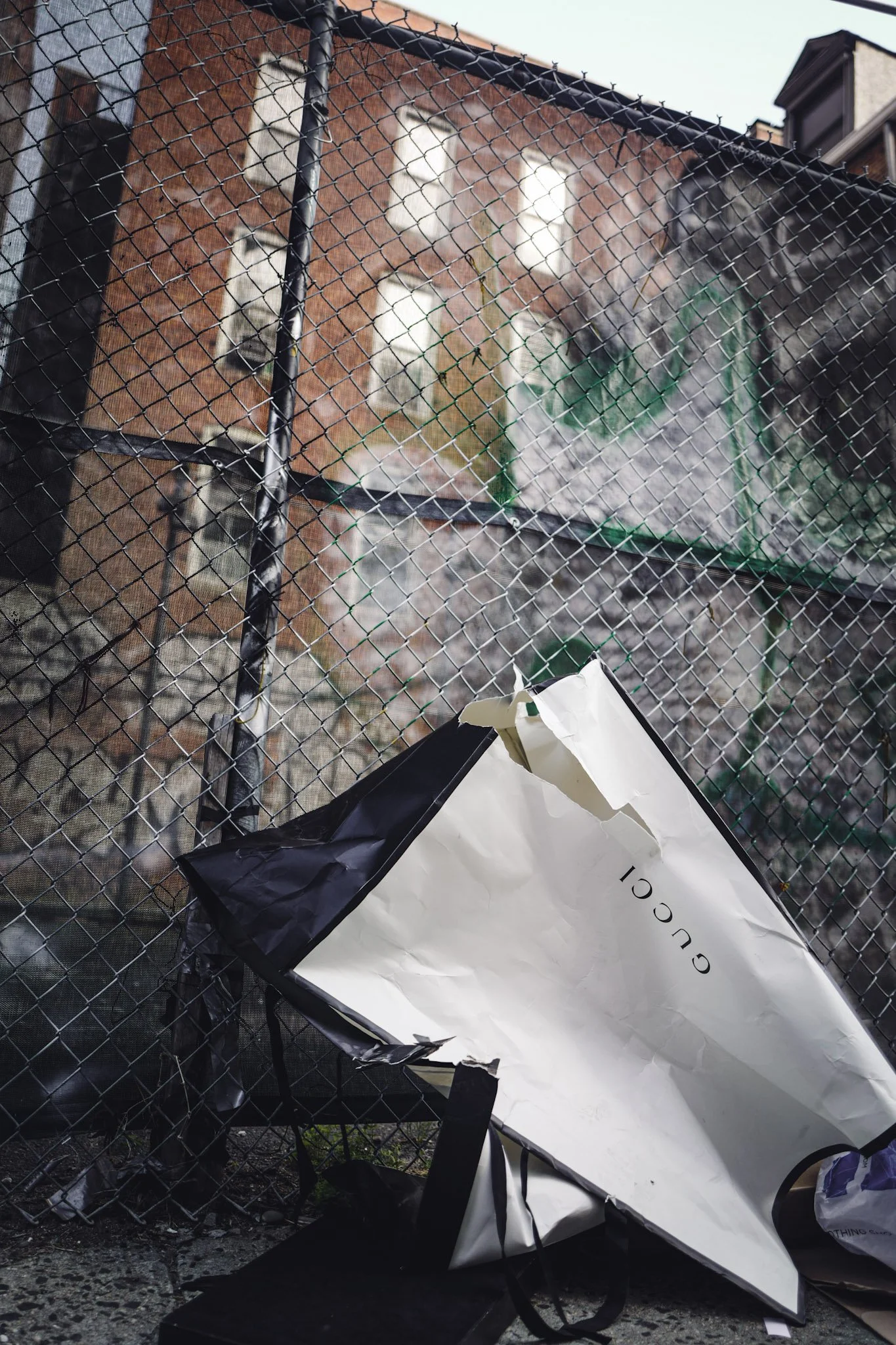 Wrecked white paper bag with black trim and the word 'GUCCI' on it, crumpled and fallen onto the ground near a chain-link fence with a damaged brick building in the background.