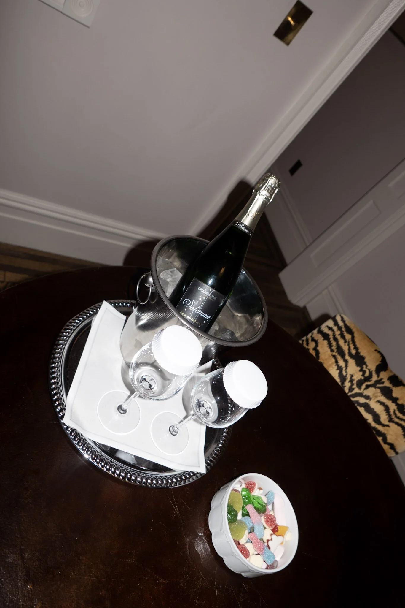 A bottle of champagne in an ice bucket, four empty glasses, and a bowl of colorful gummy candies on a dark wooden table.