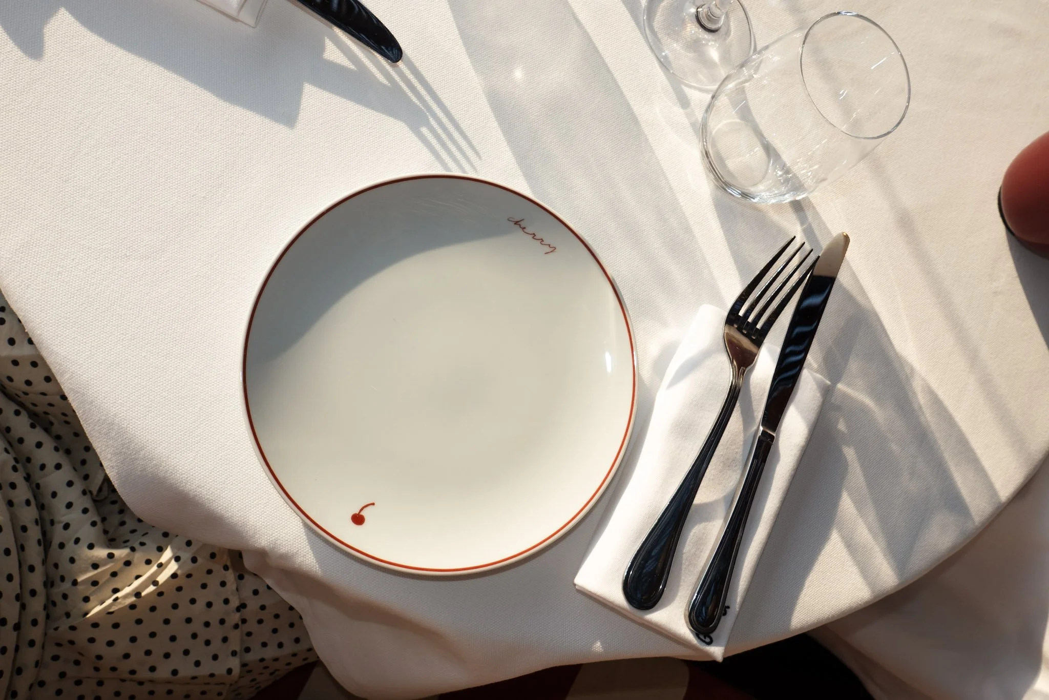 A white table setting with a plate labeled 'churron', a fork, a knife, a white cloth napkin, and an empty glass on a white tablecloth, with shadows cast by sunlight.