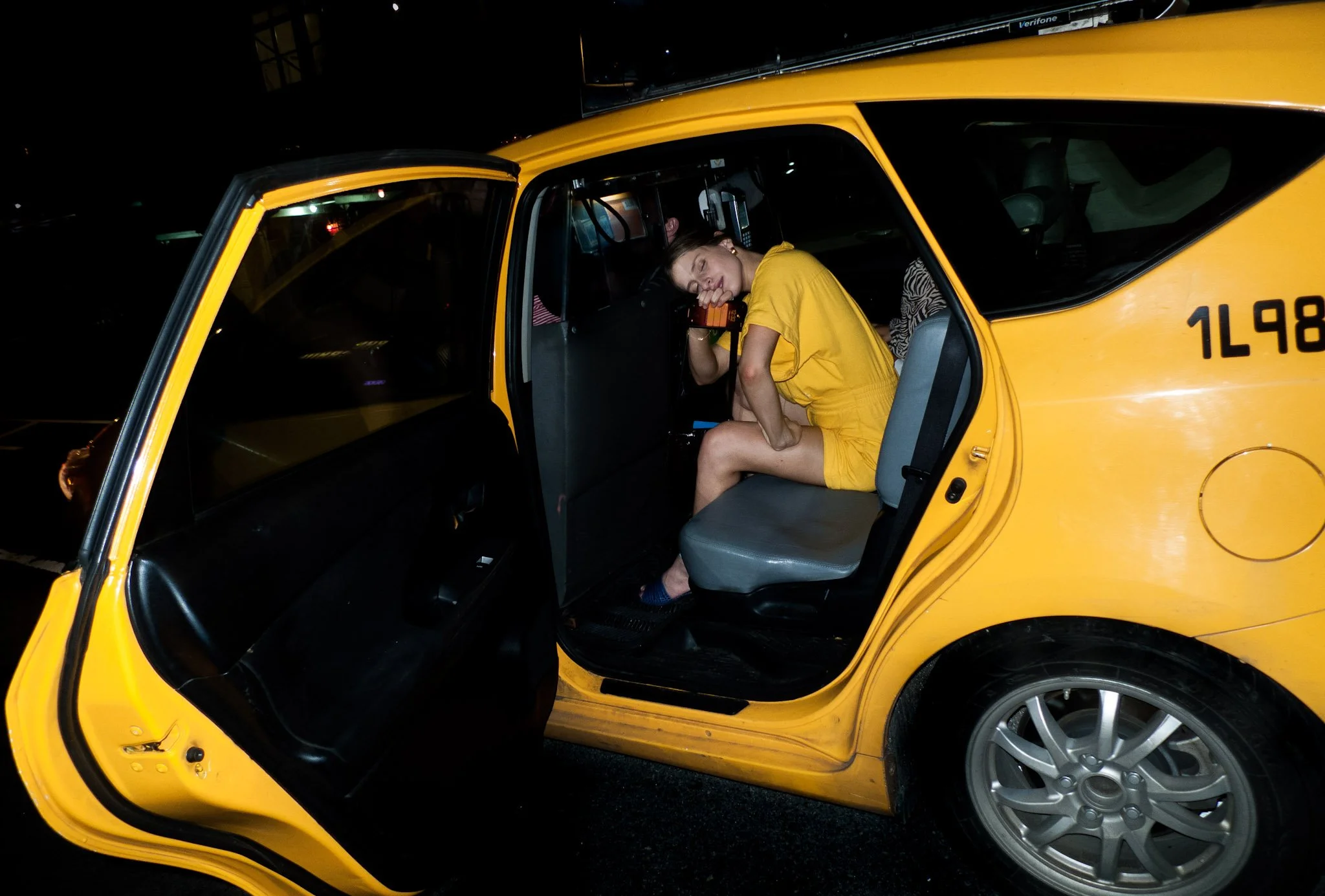 A woman in a yellow dress is sitting inside a yellow taxi at night, resting her head on the steering wheel with her eyes closed