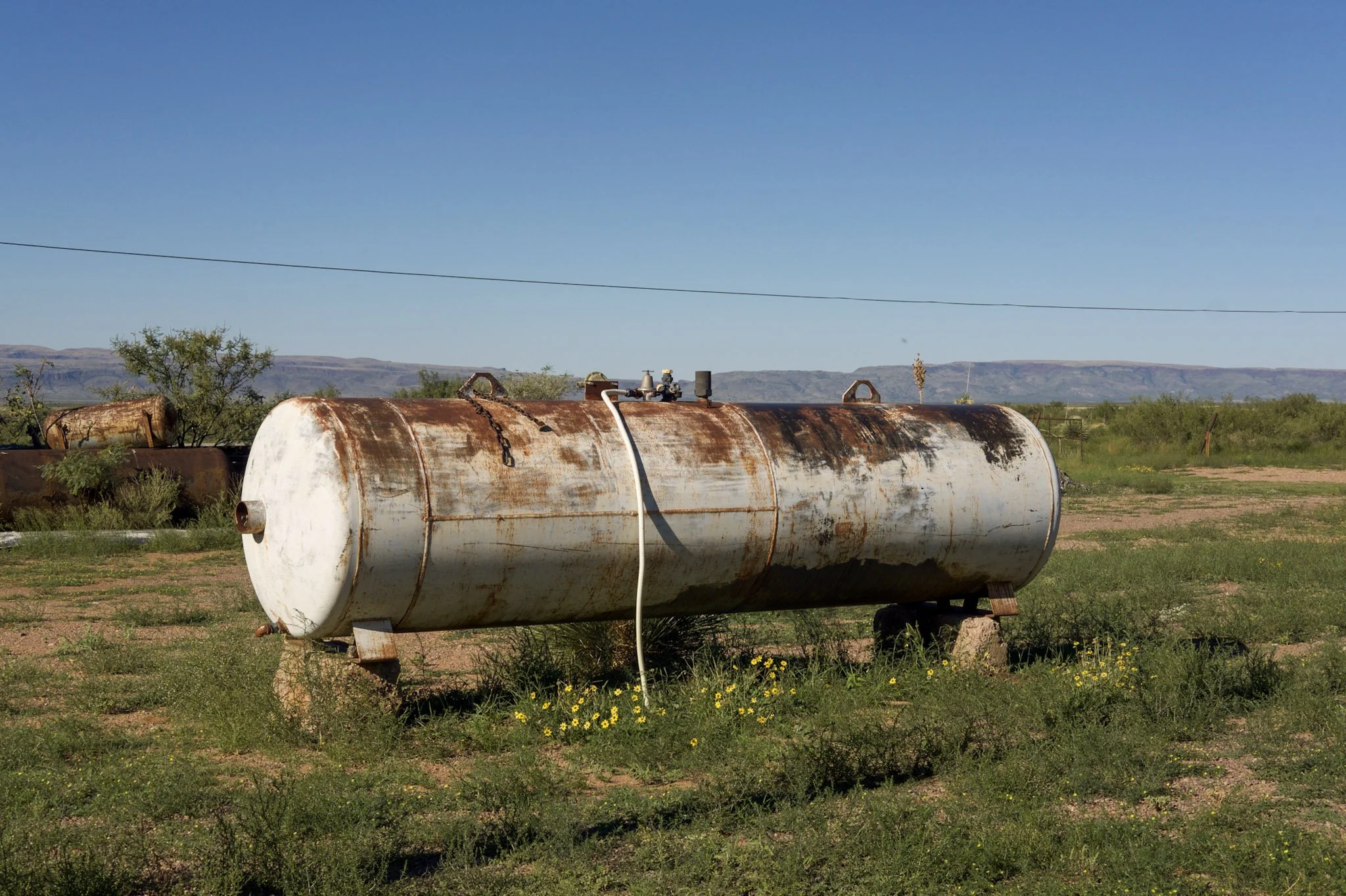 Rusty white water storage tank lying on the ground surrounded by grass and small yellow flowers, with a backdrop of green shrubs, trees, and distant mountains under a clear blue sky.
