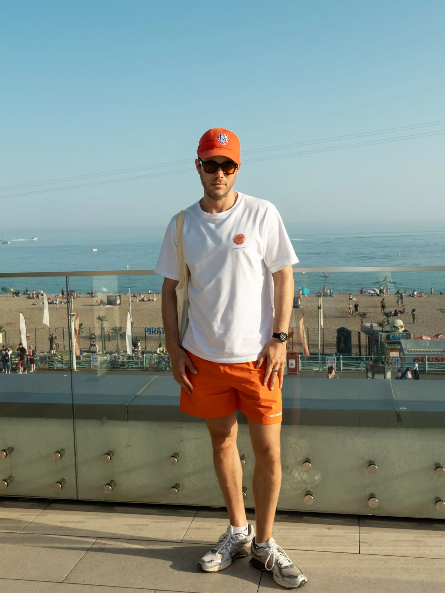 Man wearing orange cap, dark sunglasses, white t-shirt, red shorts, and sneakers standing in front of a glass railing at a beachside location with blue sky and ocean in the background.