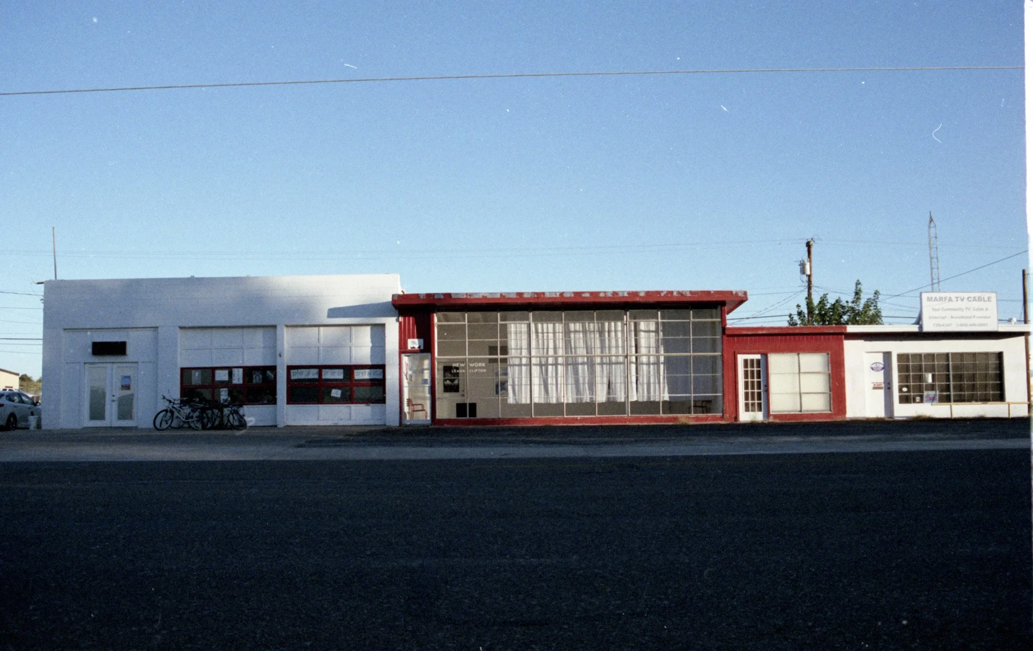 A small commercial building with white and red exterior, large glass windows, and a sign that reads 'MARFA TV CABLE.' Bikes are parked outside, and there are power lines and a clear blue sky in the background.