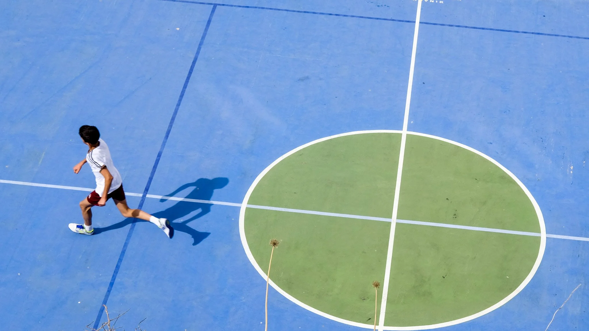 A young person in athletic clothing running on a colorful, outdoor basketball or sports court with blue and green sections, and white boundary lines.