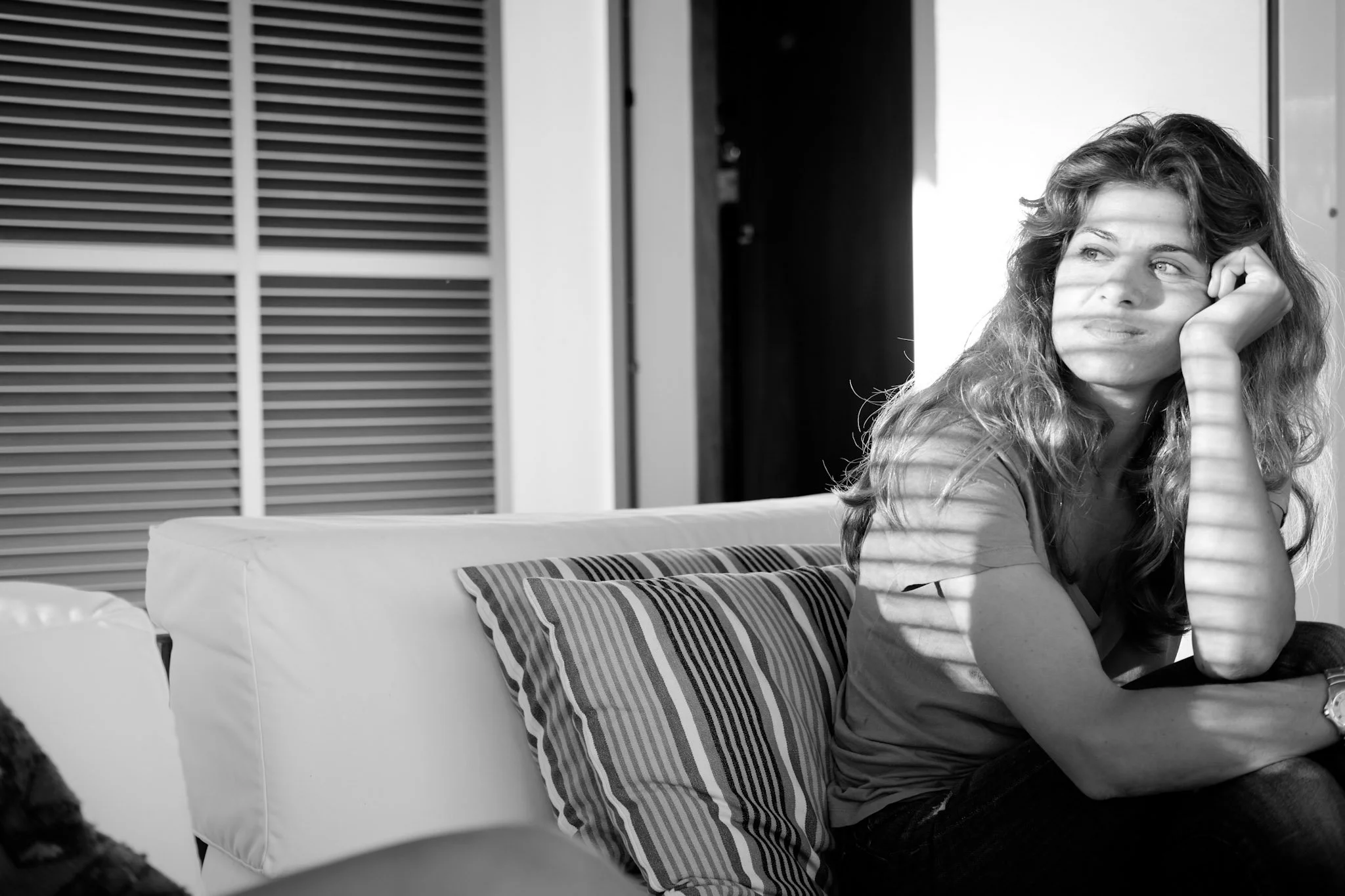 A woman with wavy hair sits on a patterned sofa, resting her chin on her hand, looking thoughtfully to the side, with sunlight casting shadows across her face.