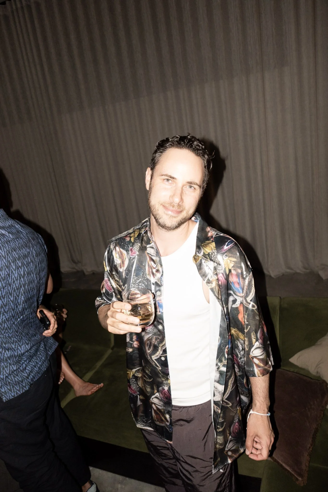 A man in a floral shirt holding a glass of white wine at a party.