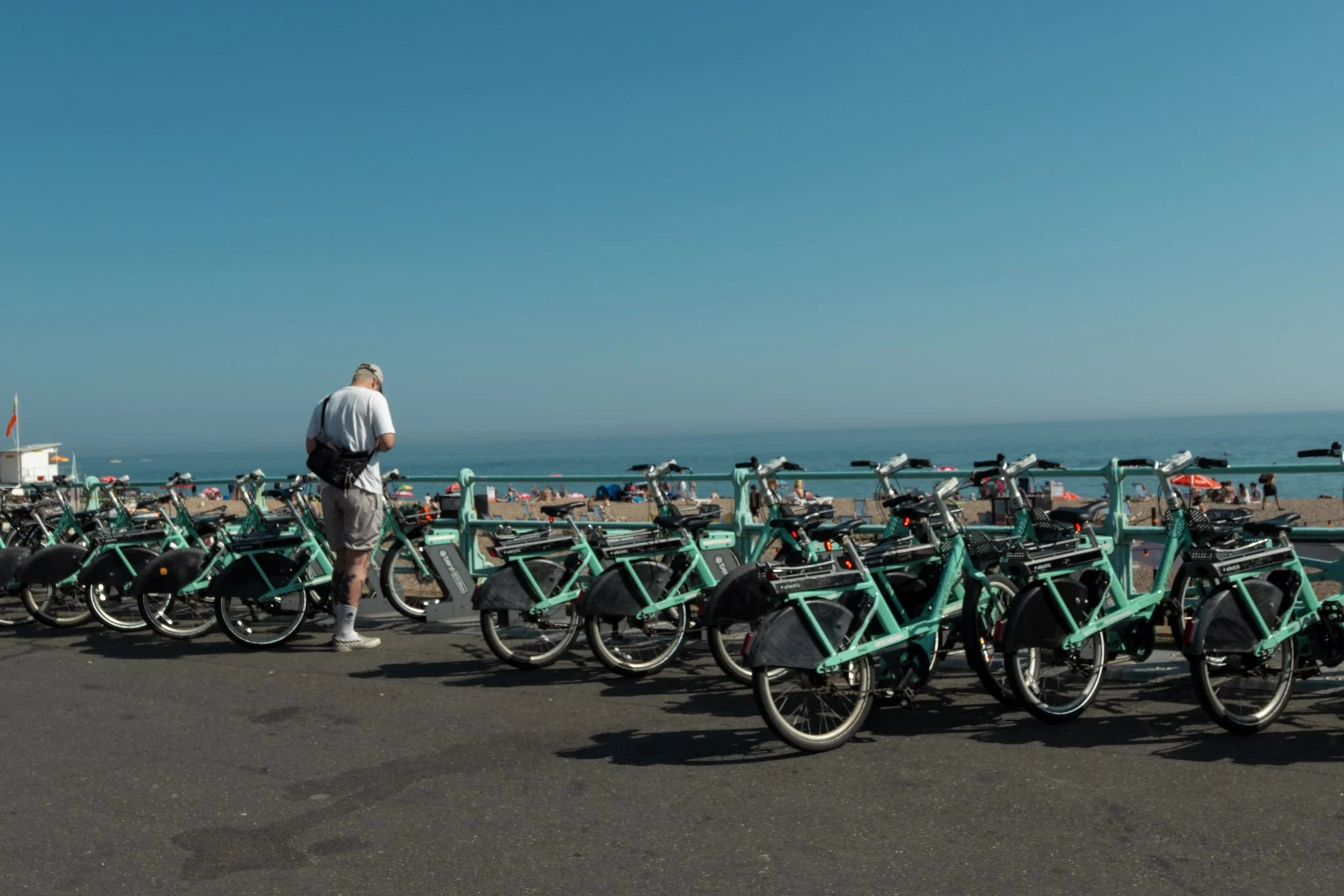 A man standing among a row of turquoise electric scooters at a beachside area with people and umbrellas in the background, and the ocean and clear blue sky beyond.