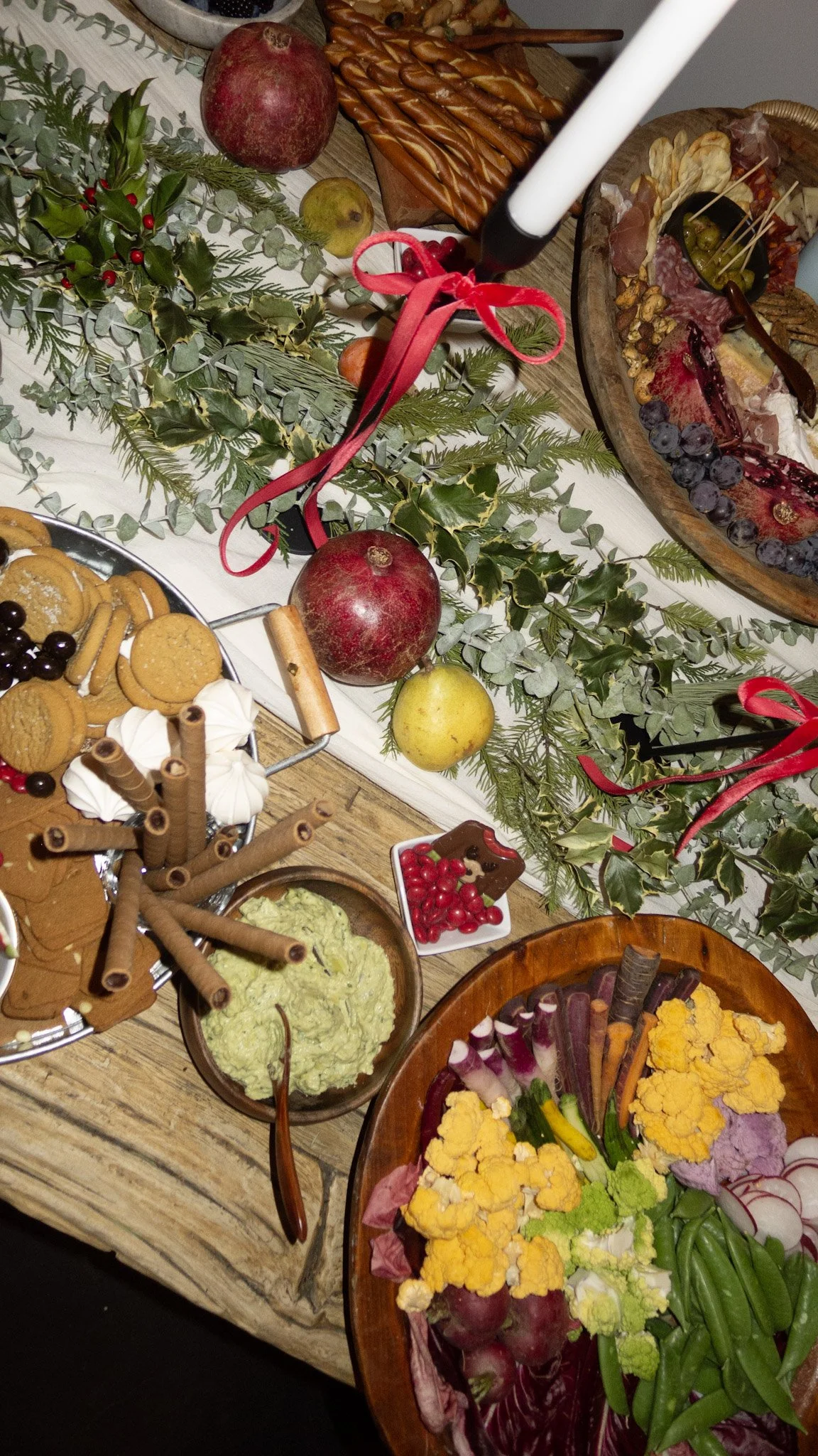A holiday buffet table decorated with greenery, holly, red ribbons, and a candle, featuring an assortment of foods including cheese, crackers, grapes, vegetables, and festive dishes.