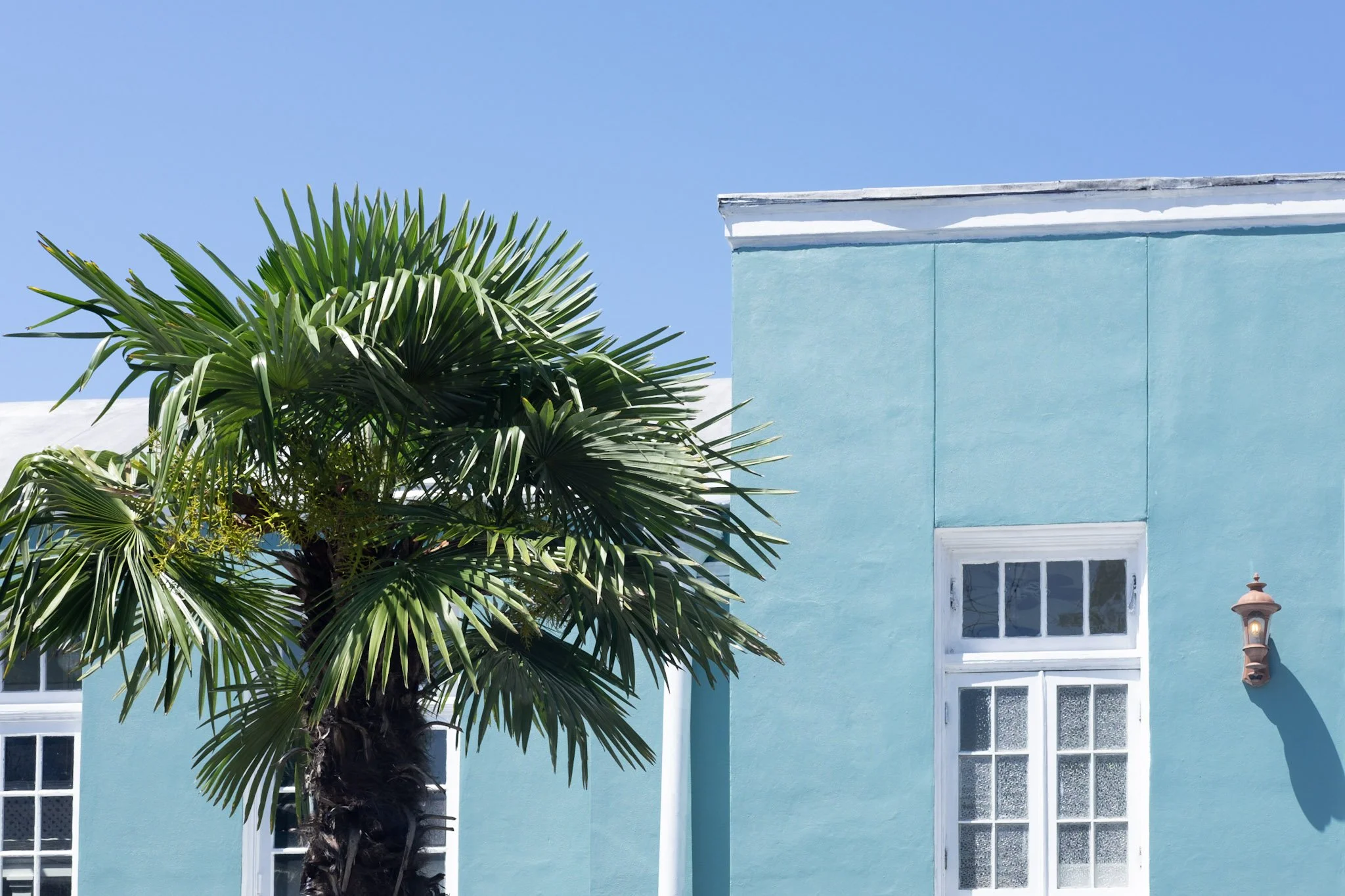 A turquoise building with white trim, a window with grid panes, and a small lantern on the wall. There is a green palm tree with long, pointed fronds in front of the building, and a clear blue sky in the background.
