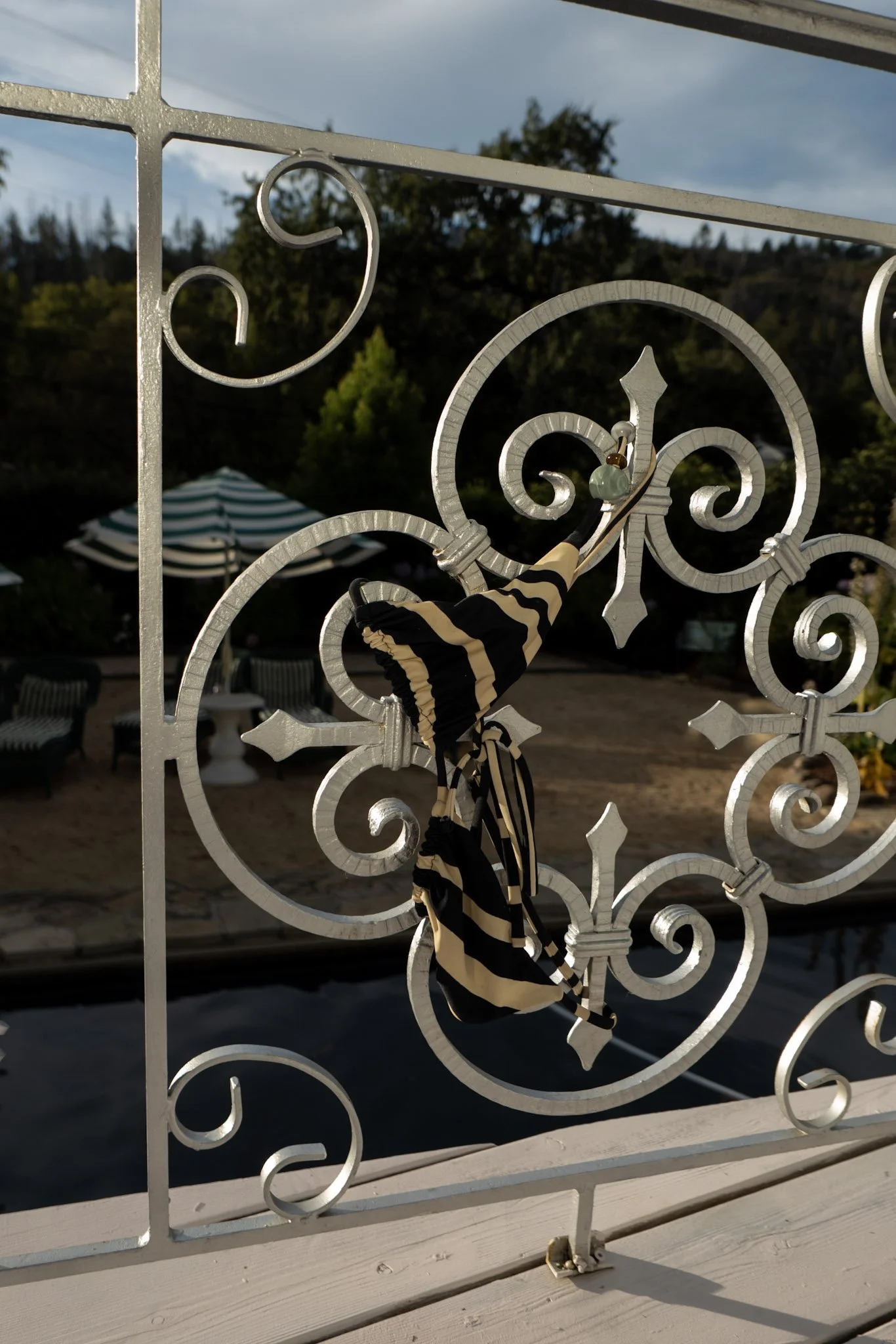 Decorative white metal railing with intricate swirl patterns, with a black and beige striped silk scarf hanging from it. In the background, there are trees, a cloudy sky, and striped umbrellas likely on a terrace or outdoor café area.