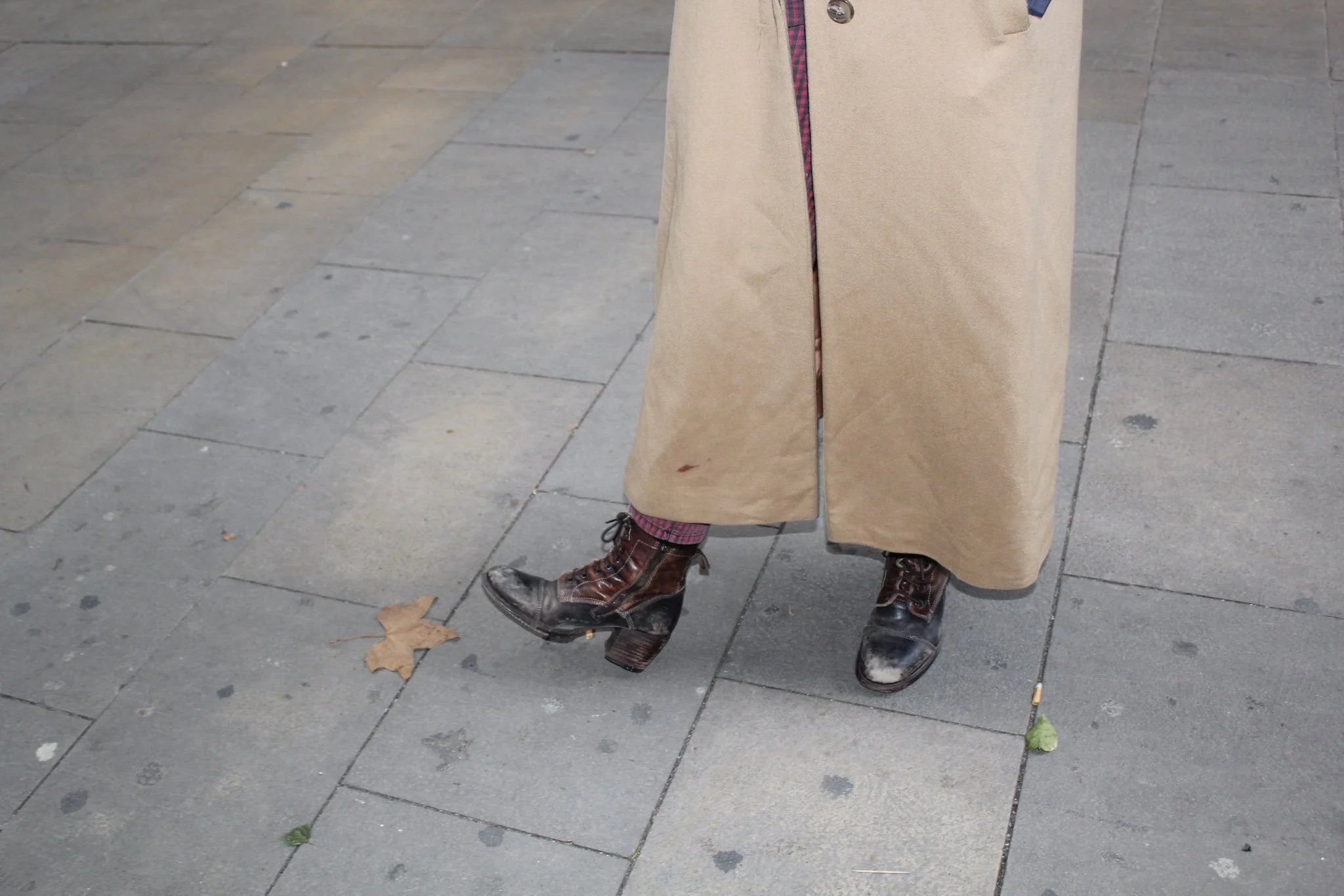 Person standing on a sidewalk wearing a long beige coat, patterned pants, and brown leather boots, holding a green leaf on a stick.