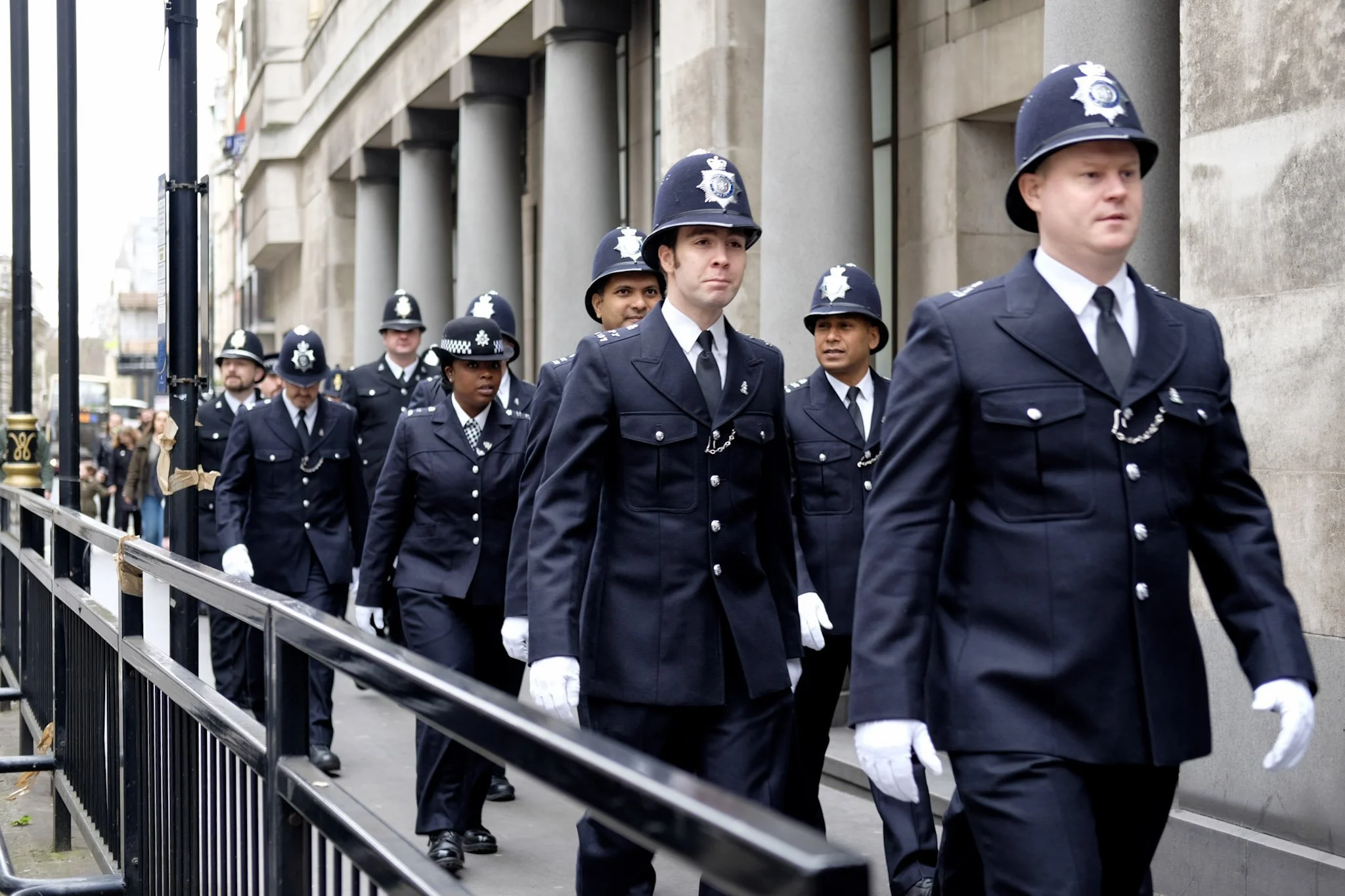 A group of police officers in formal uniform march in a parade along a city street, with buildings in the background.