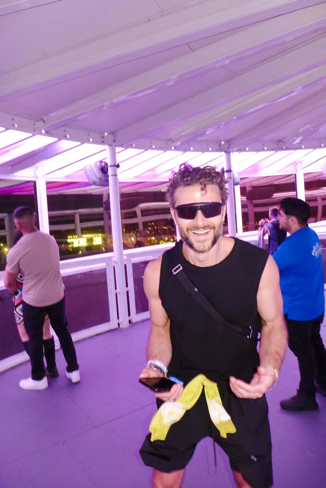 Smiling man wearing sunglasses holding a phone with a yellow jacket tied around his waist, standing under purple lighting on a rooftop.