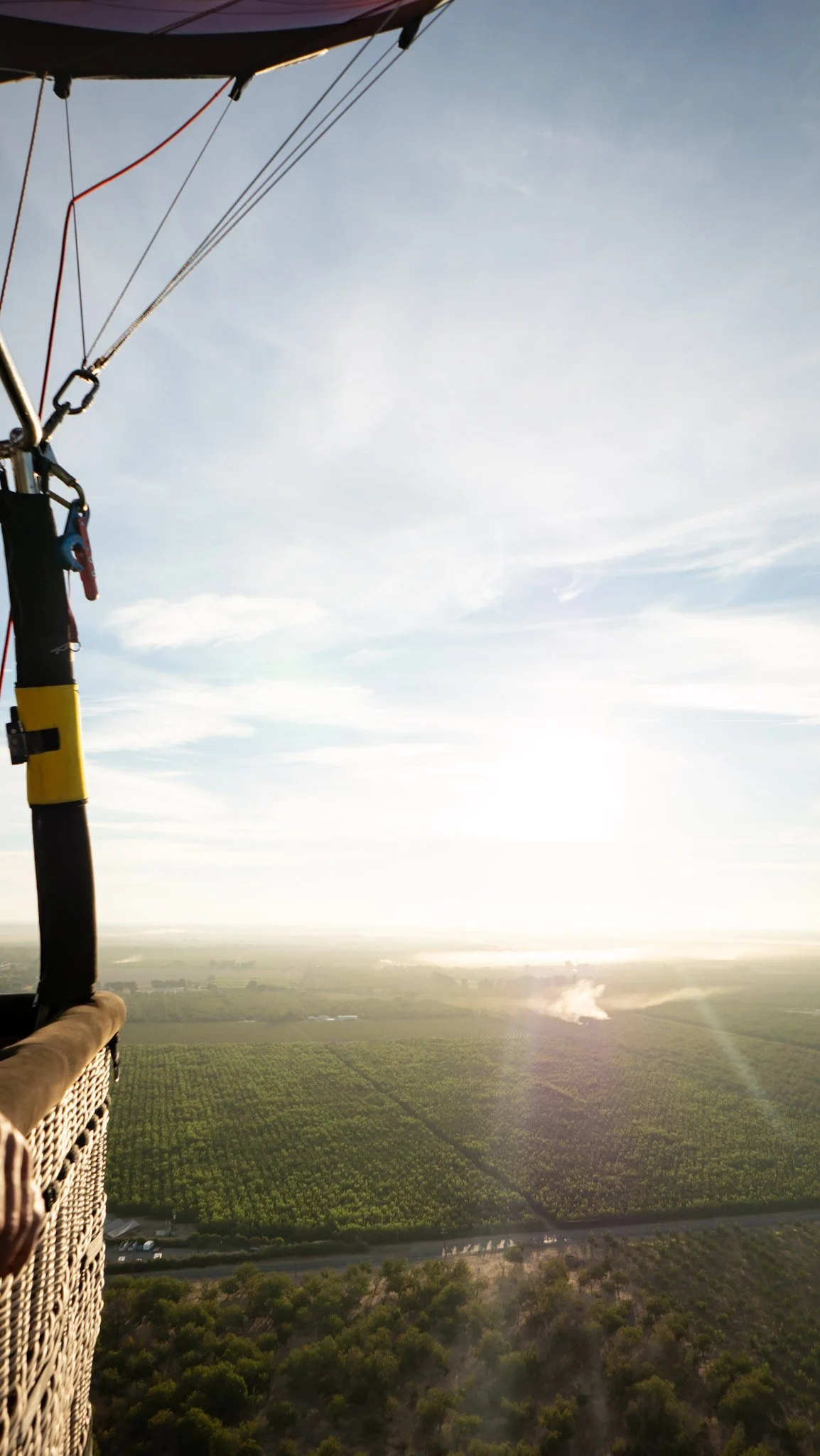 View from a hot air balloon flying over fields and trees, with a clear sky in the background during sunset or sunrise.