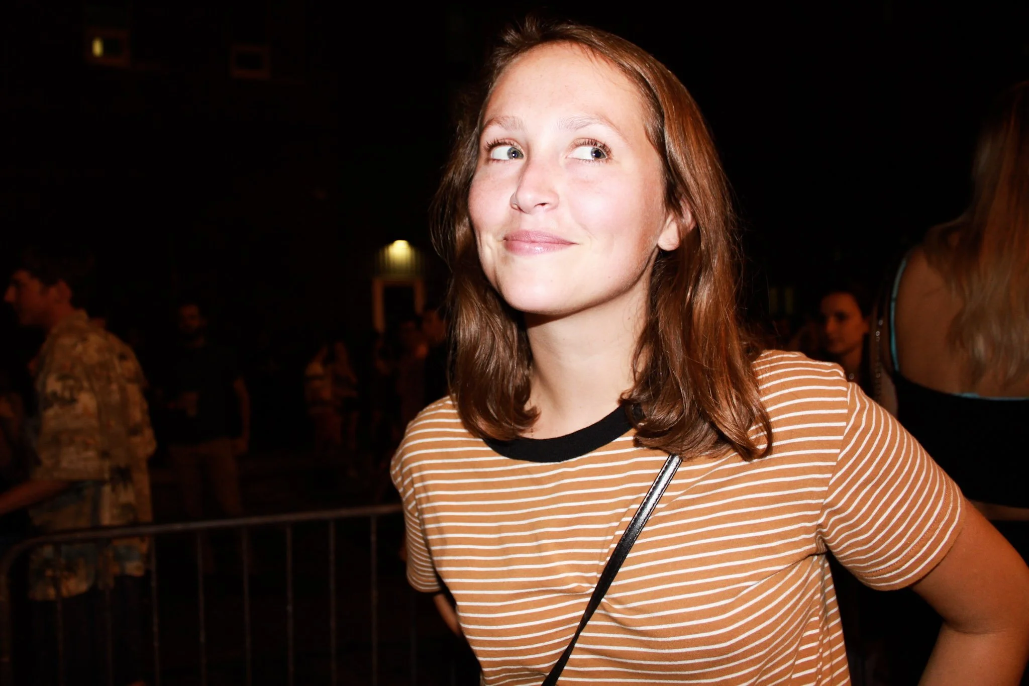 A young woman with red hair, wearing a brown and white striped shirt, smiling and looking to her right, in a dark indoor setting with other people in the background.